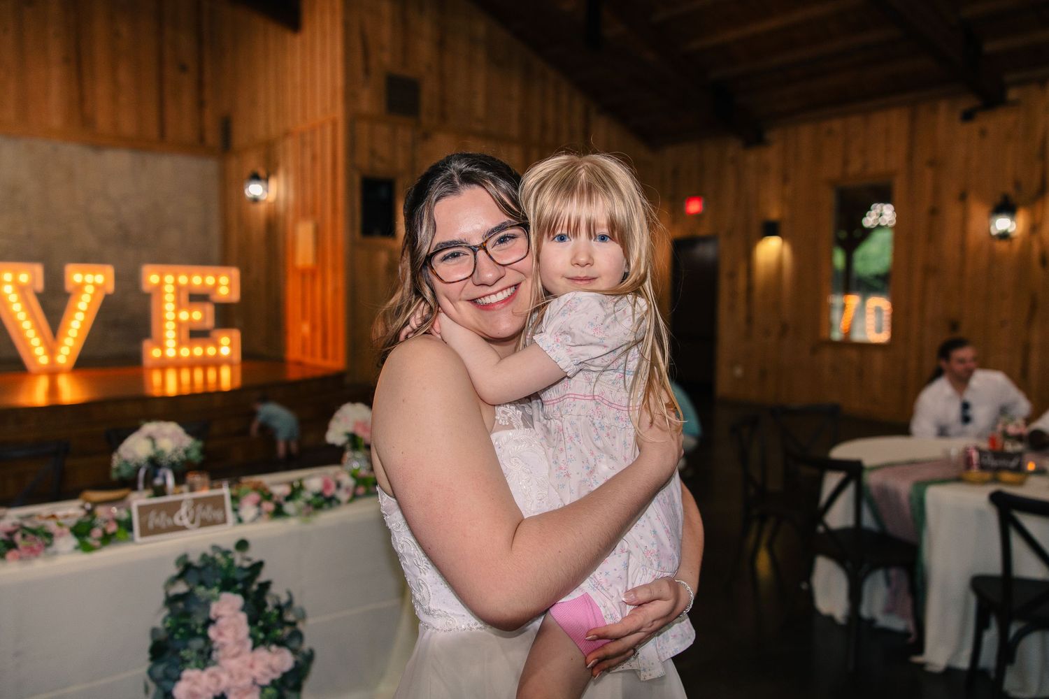 Smiling bride in a white lace gown holds a little girl in a floral dress at a barn wedding reception with marquee lights.