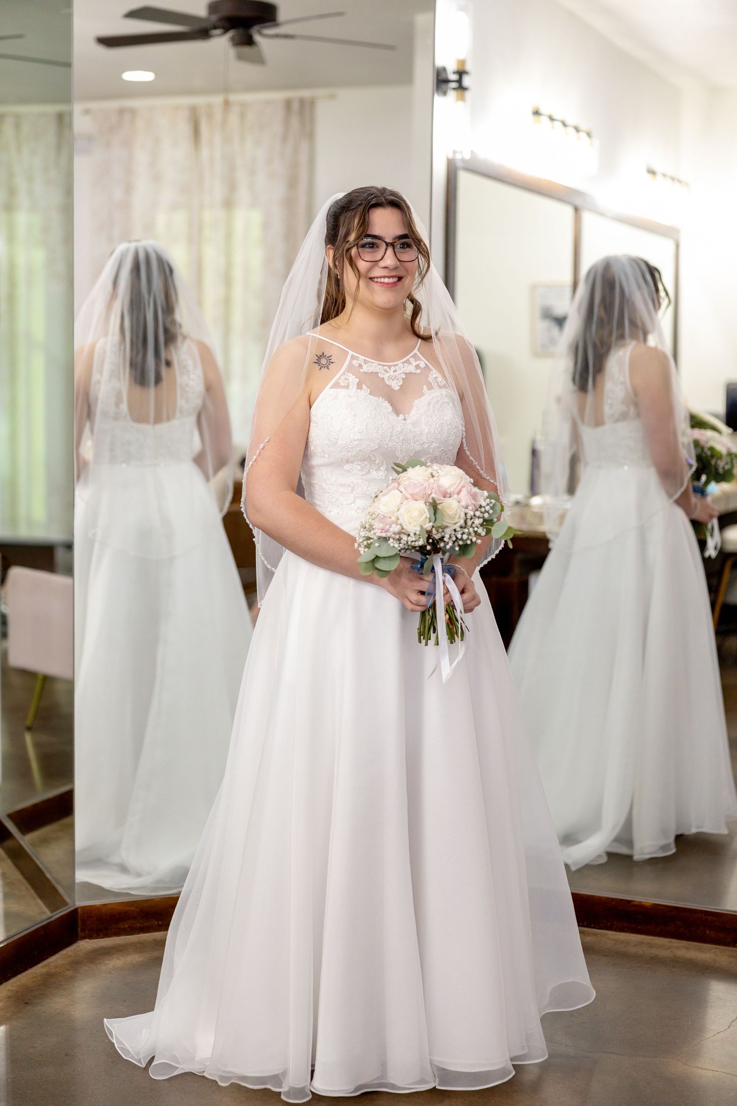 Smiling bride in glasses wearing a lace-bodice wedding gown and veil holds a pink bouquet in front of mirrors.