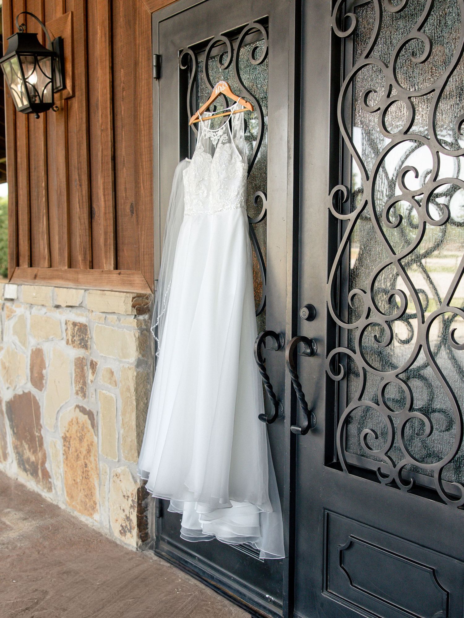 White lace wedding dress with a flowing tulle skirt hanging on a wooden hanger against an ornate wrought iron door and stone wall.
