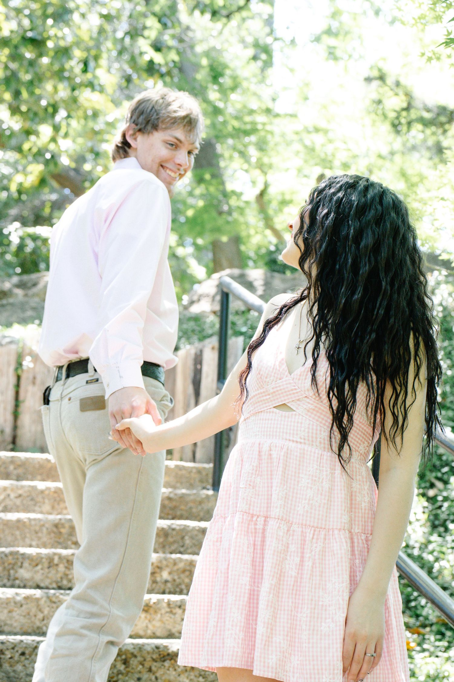 Happy couple holding hands on outdoor stone staircase surrounded by lush green trees.