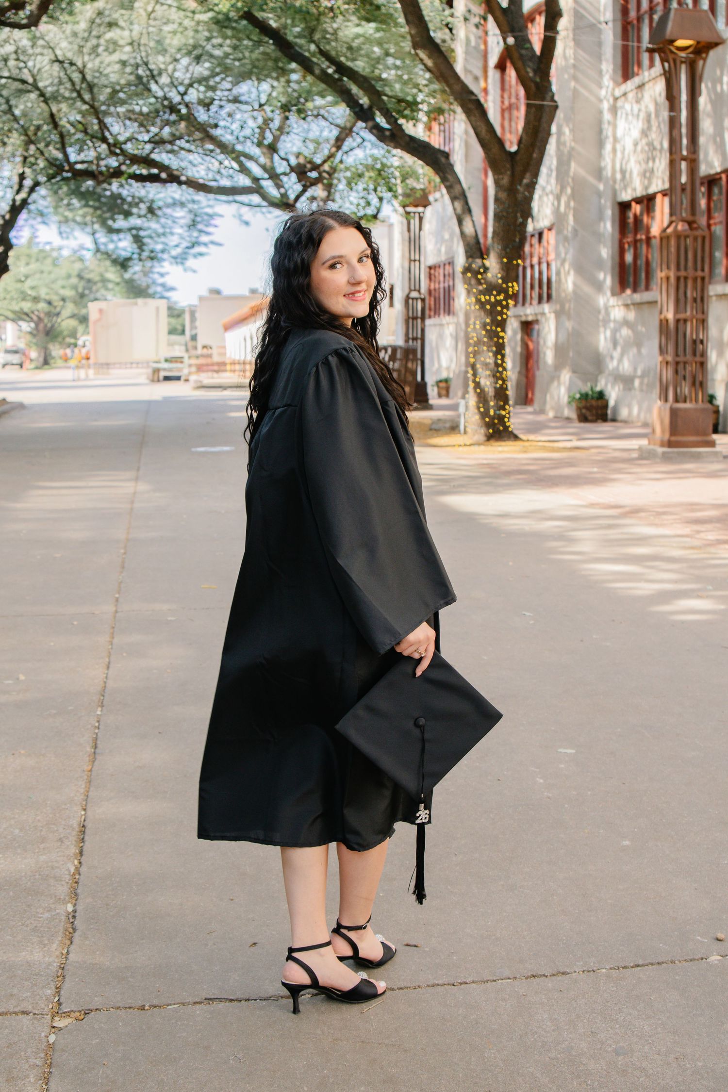 Graduate in black gown holding cap while walking on tree-lined sidewalk, glancing back over shoulder.
