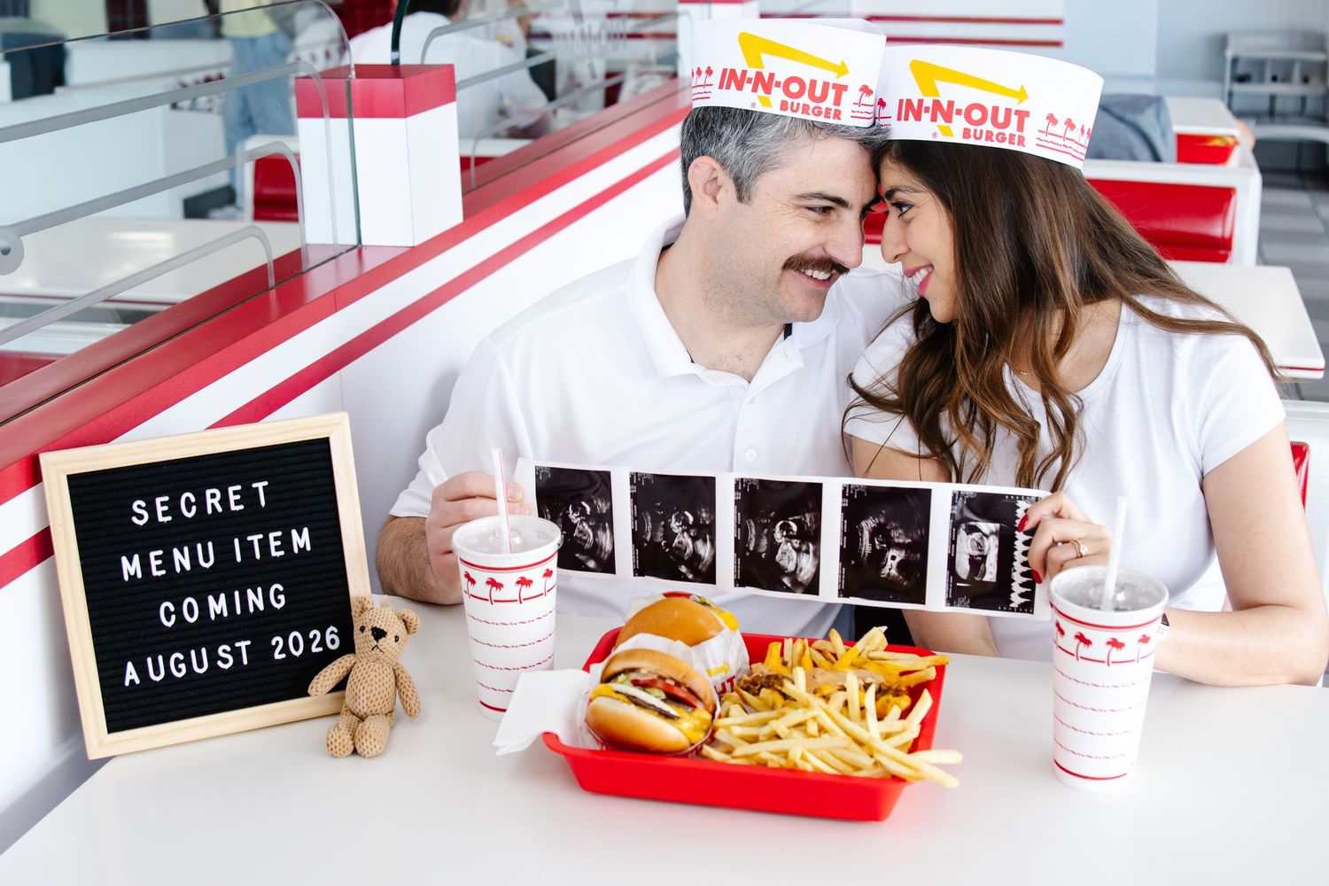 Couple at In-N-Out Burger holding ultrasound strip next to pregnancy announcement letter board and food.