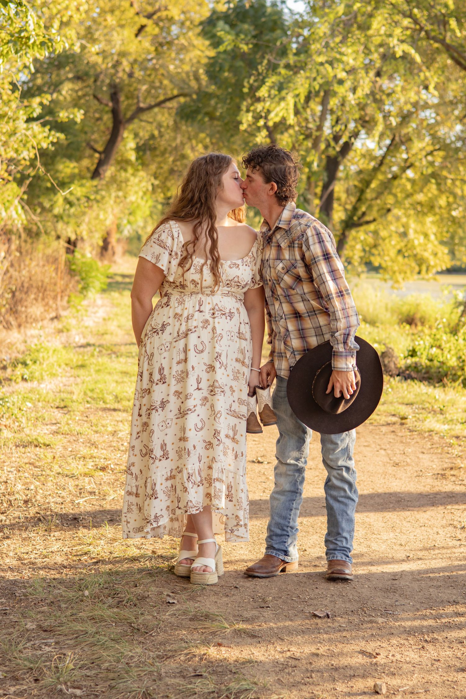 Couple kissing outdoors in golden light, woman in floral dress, man in plaid shirt holding cowboy hat.