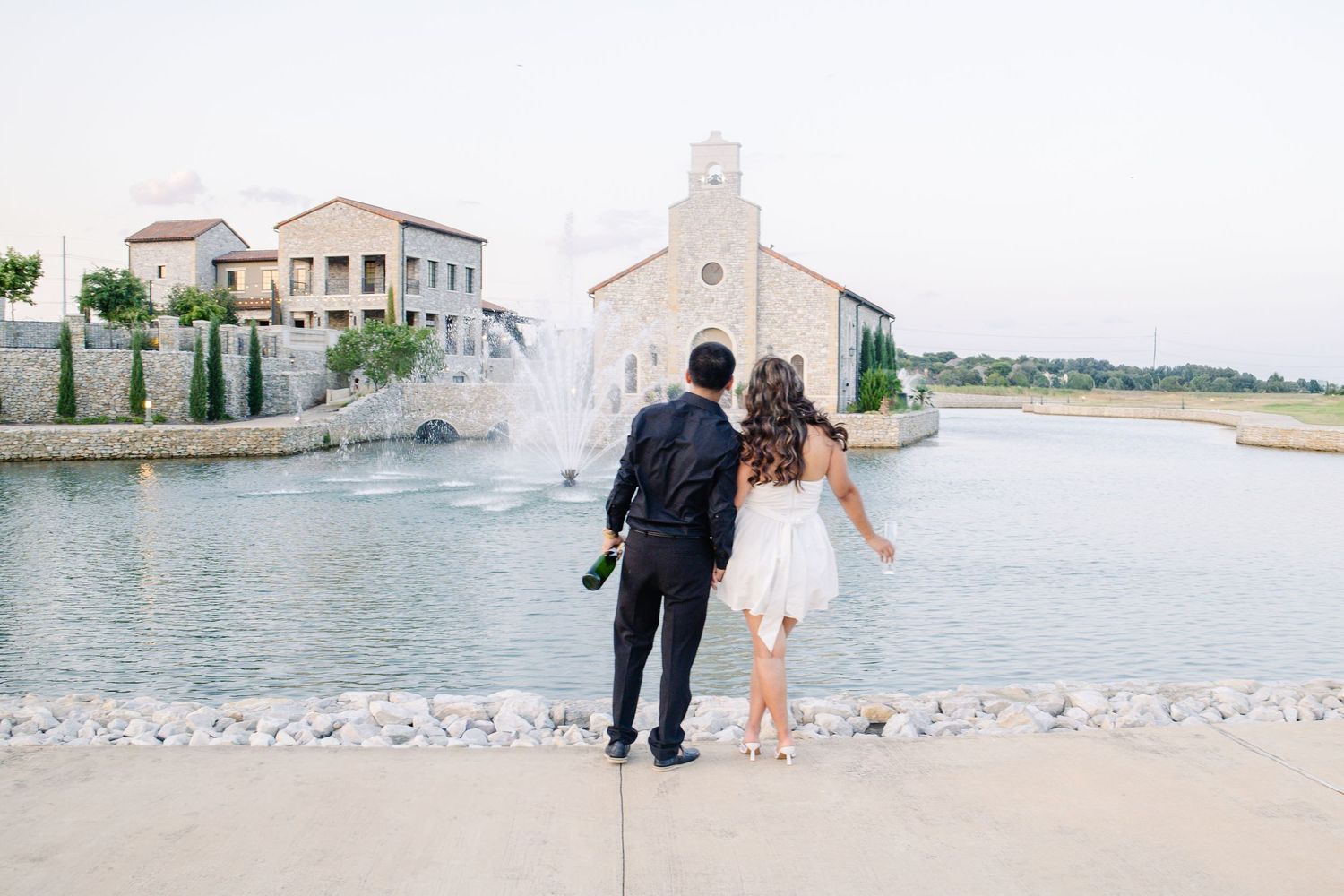 Couple holding champagne admiring fountain and chapel venue by lake at dusk.