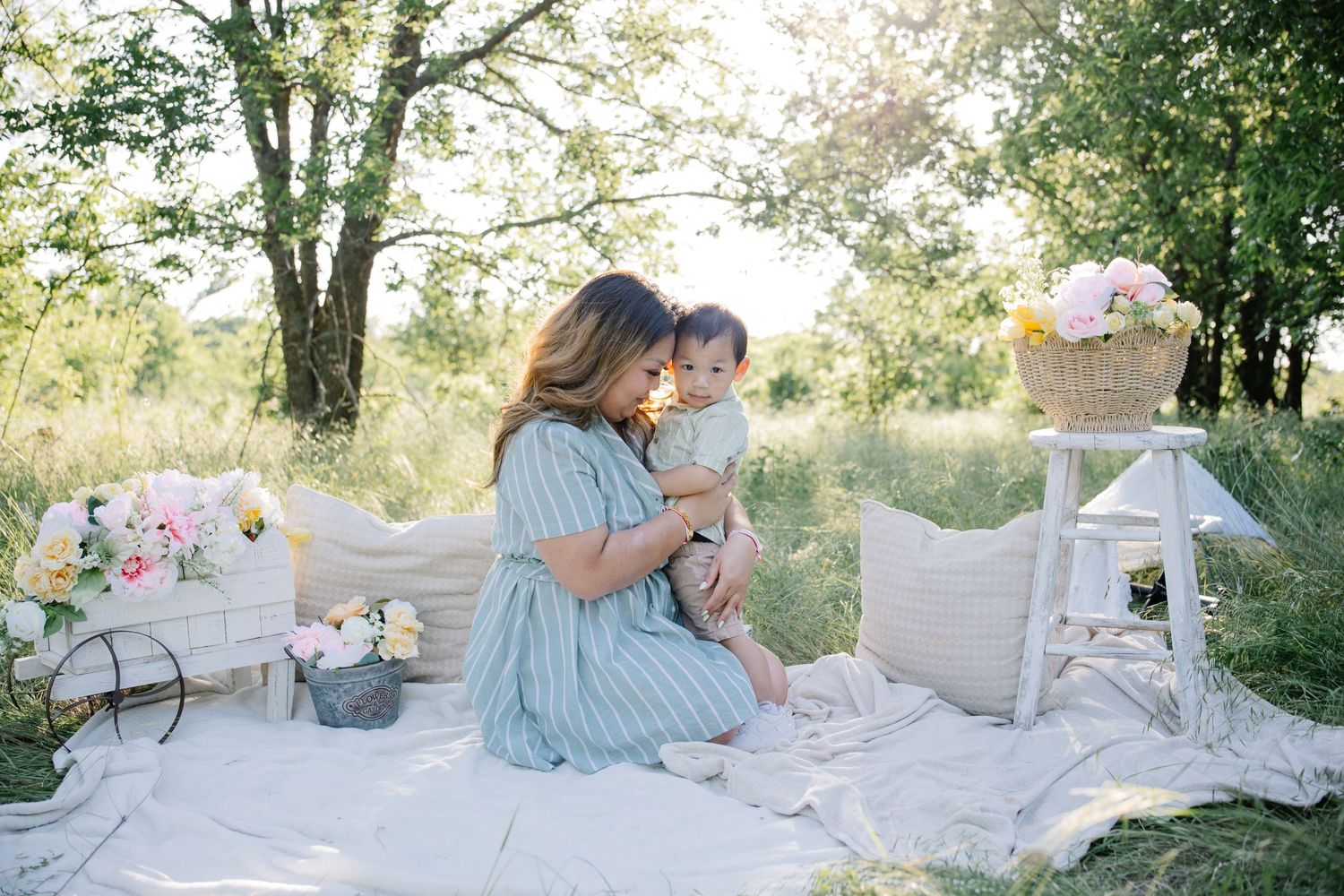 A mother in a blue dress sits on a white blanket outdoors, lovingly holding her baby amid floral decorations.