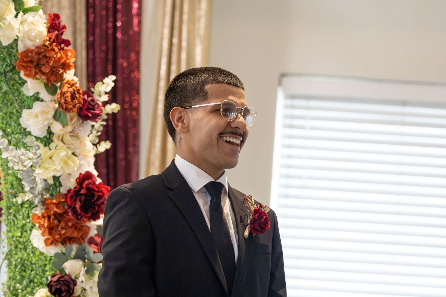 Smiling groom in black suit and glasses stands beside floral arrangement of red and white flowers at wedding ceremony.