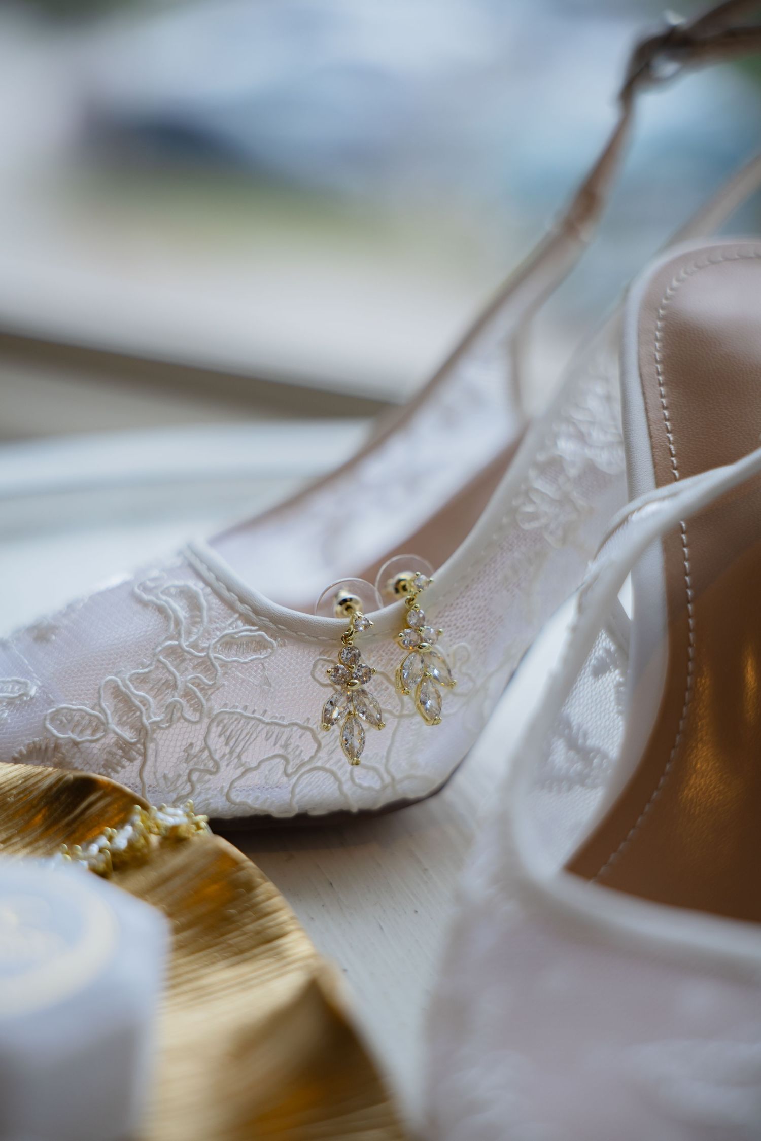 White lace bridal heels with gold crystal drop earrings displayed on a golden tray for a wedding day.