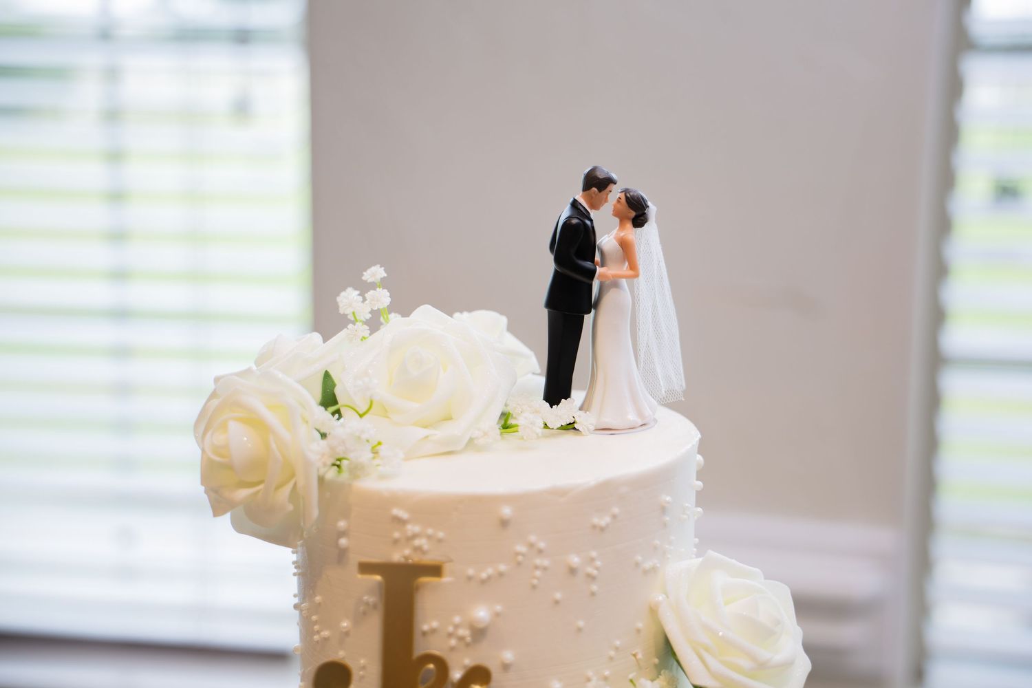 A two-tier white wedding cake topped with bride and groom figurines and decorated with white roses.