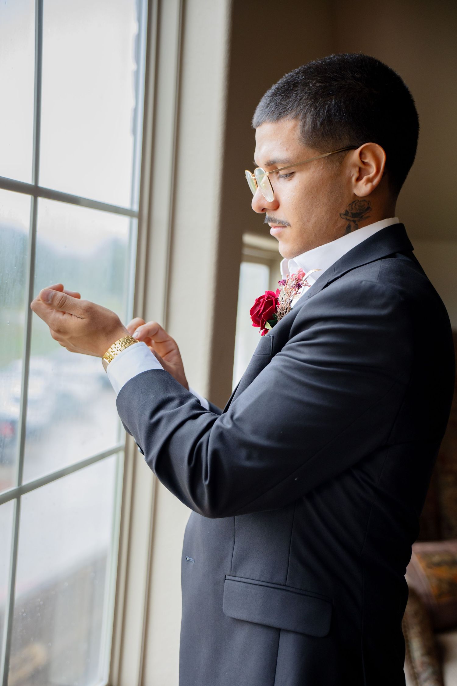 Dapper groom in navy suit with red boutonniere adjusts his gold cufflinks by a sunlit window.