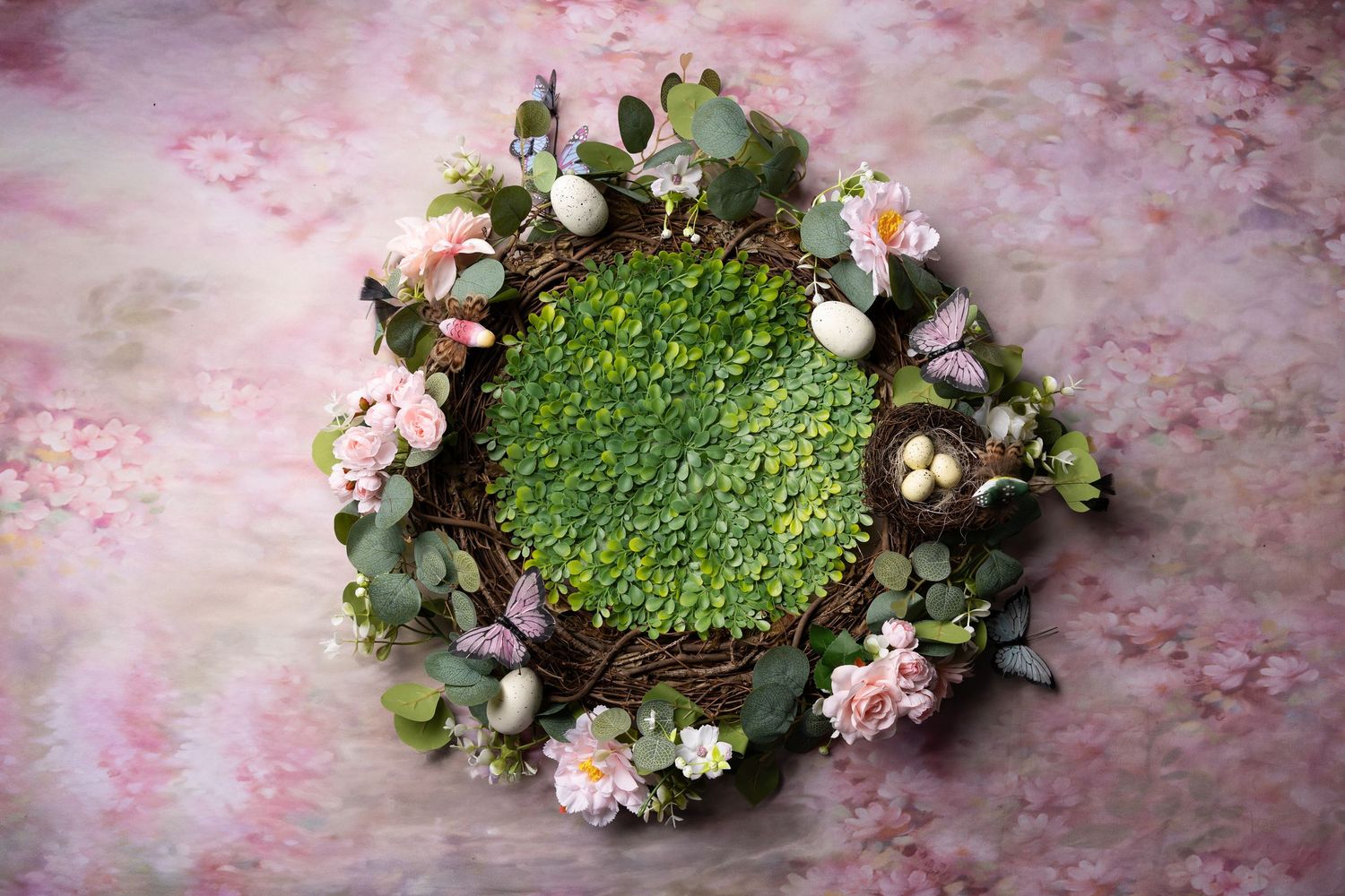 A wicker nest filled with green moss and Easter eggs, surrounded by pink flowers and eucalyptus on a floral backdrop.