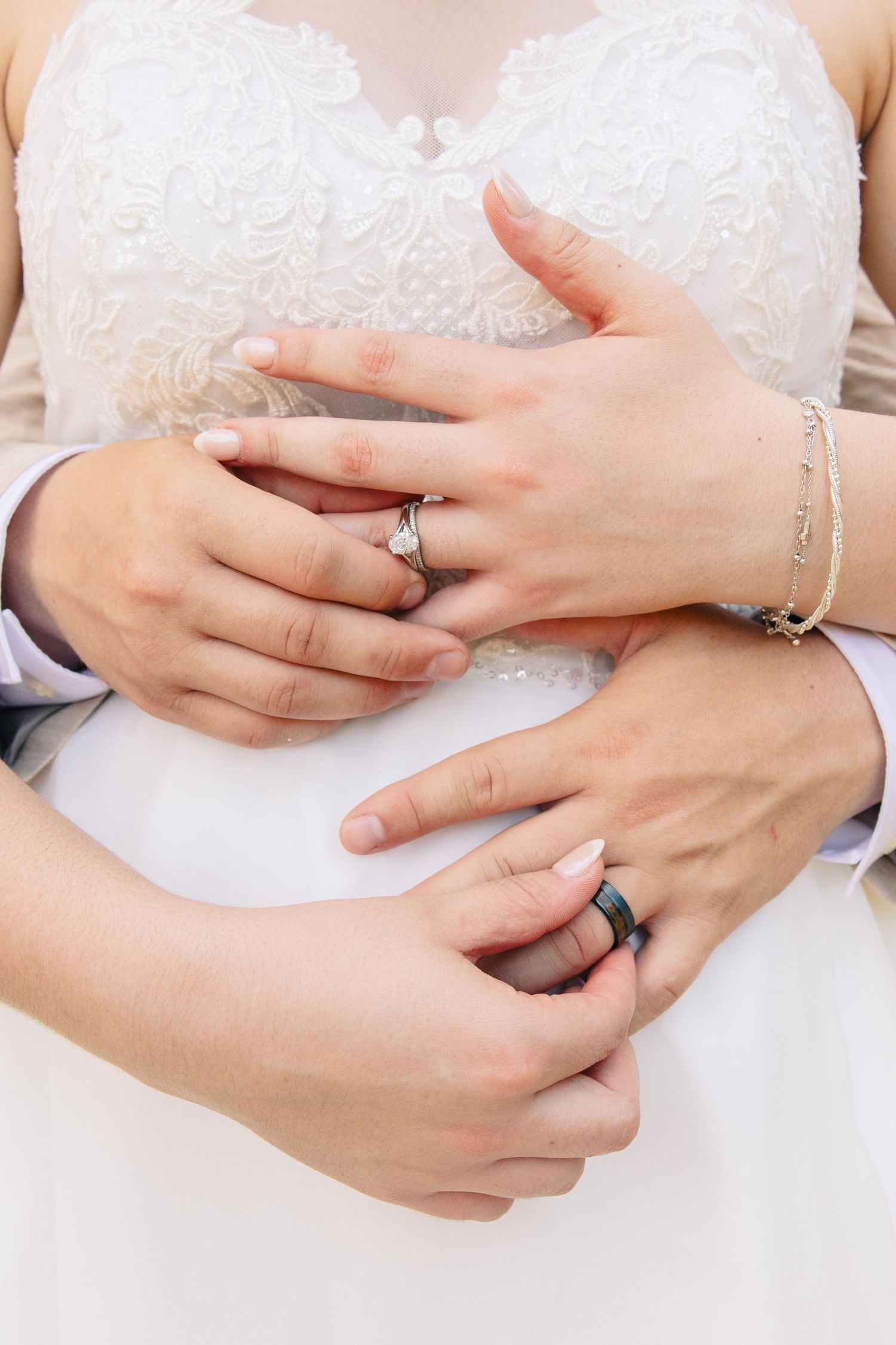 Close-up of a bride and groom's hands layered over a lace wedding dress, showing a silver engagement ring and dark wedding band.