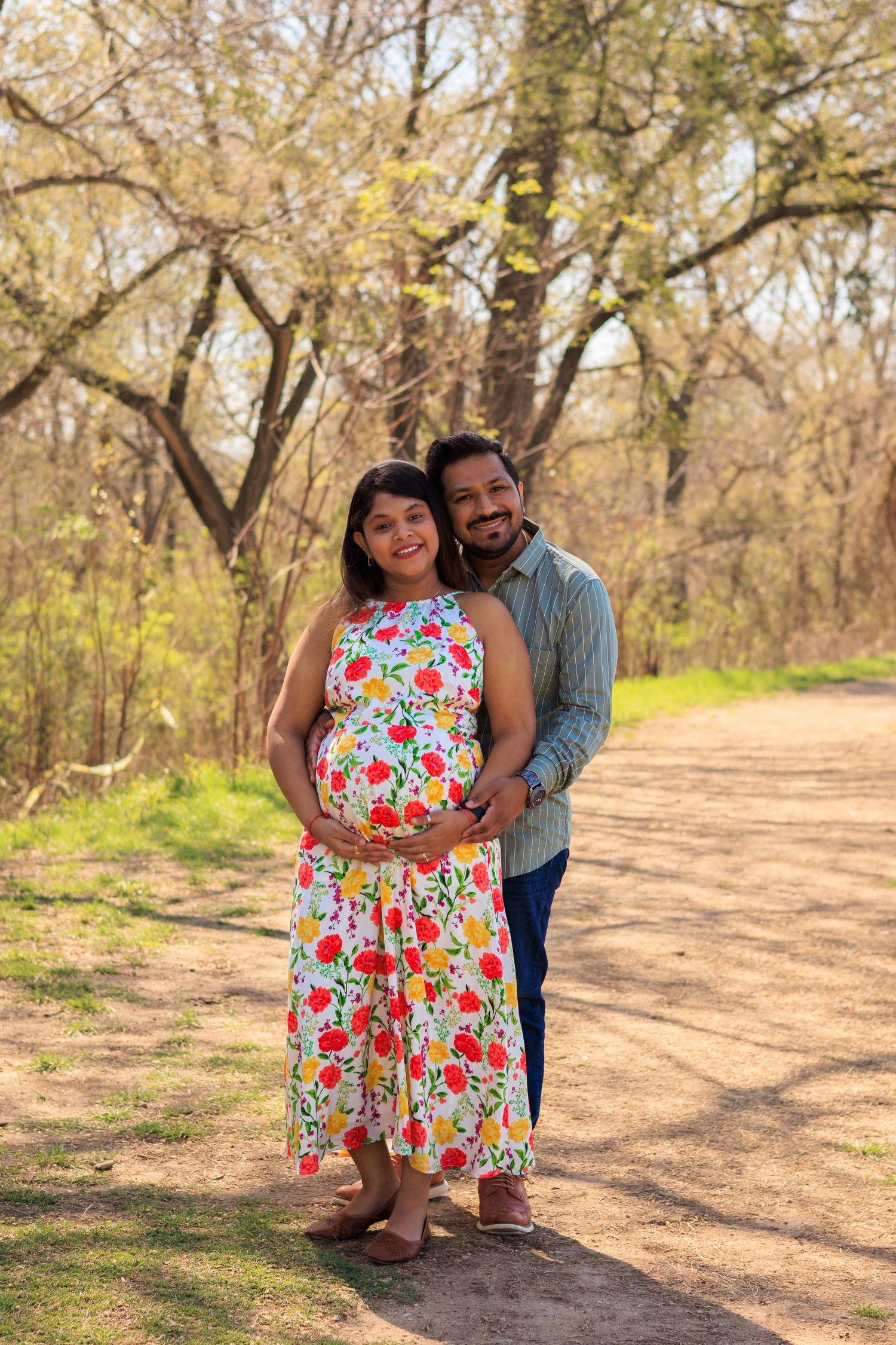 Happy expecting couple embracing on a sunlit wooded path during a spring maternity photoshoot.