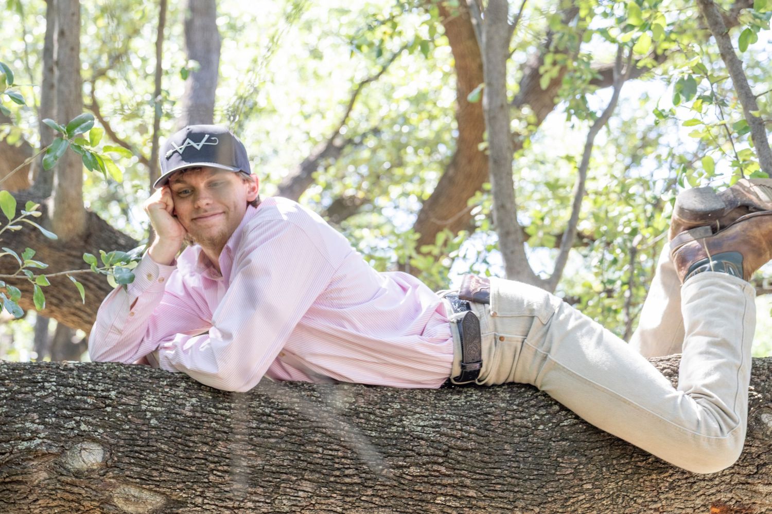 Young man in pink shirt and cap relaxes on fallen tree trunk in sunny wooded setting.