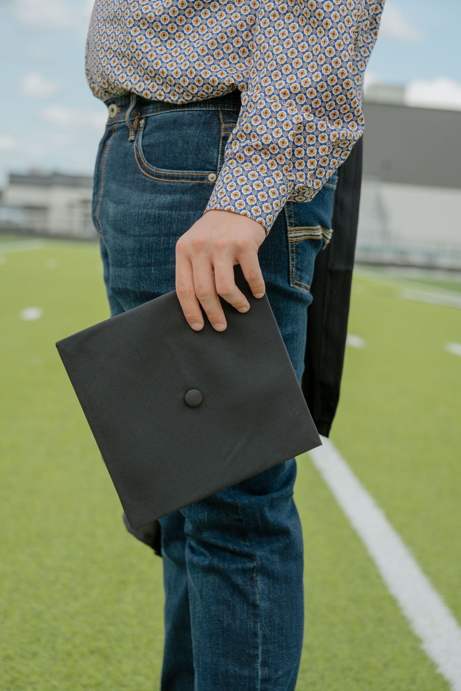 Close-up of graduate holding black mortarboard cap while wearing patterned shirt and jeans on a field.