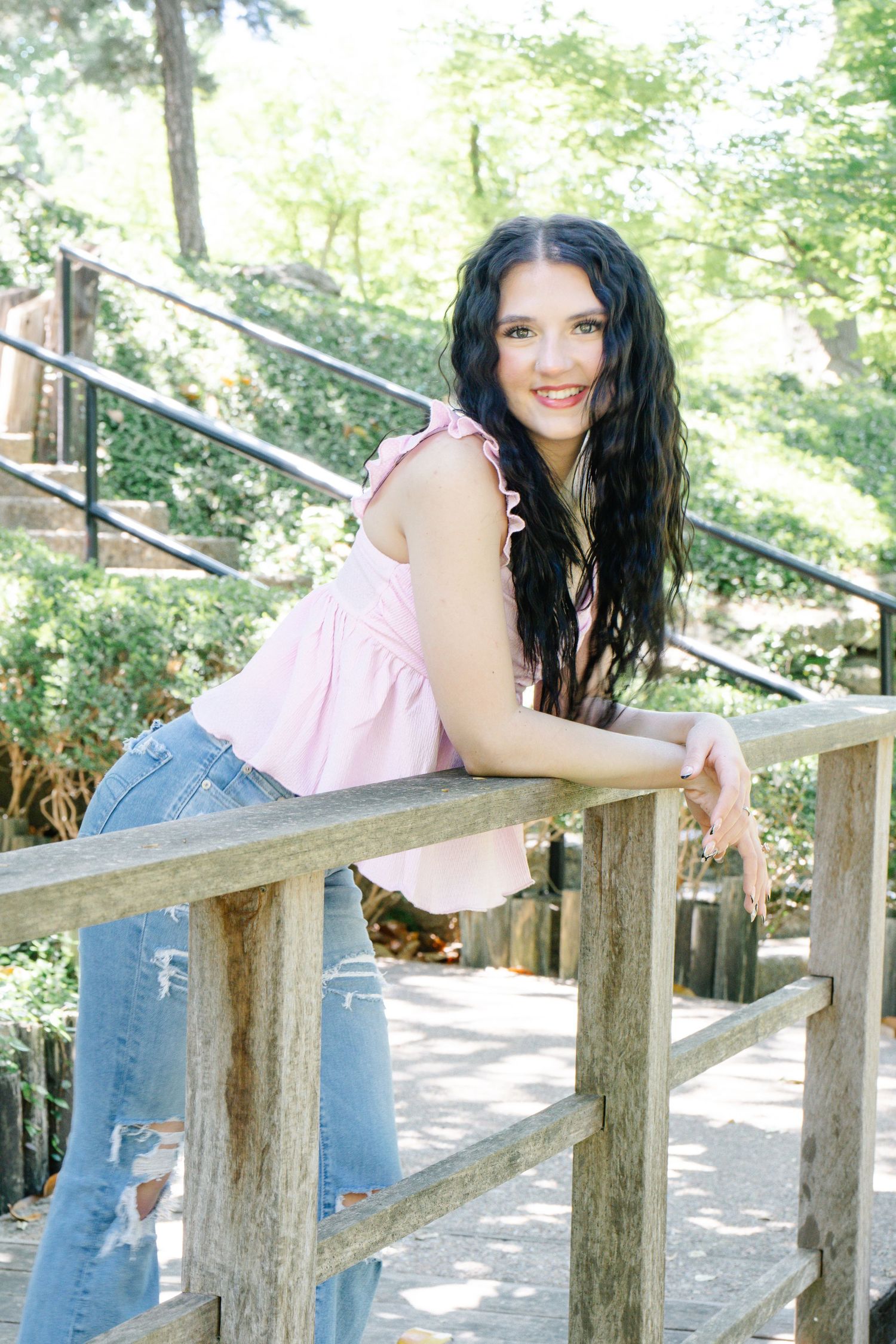 A young brunette woman in a pink top and ripped jeans poses on a wooden fence outdoors.