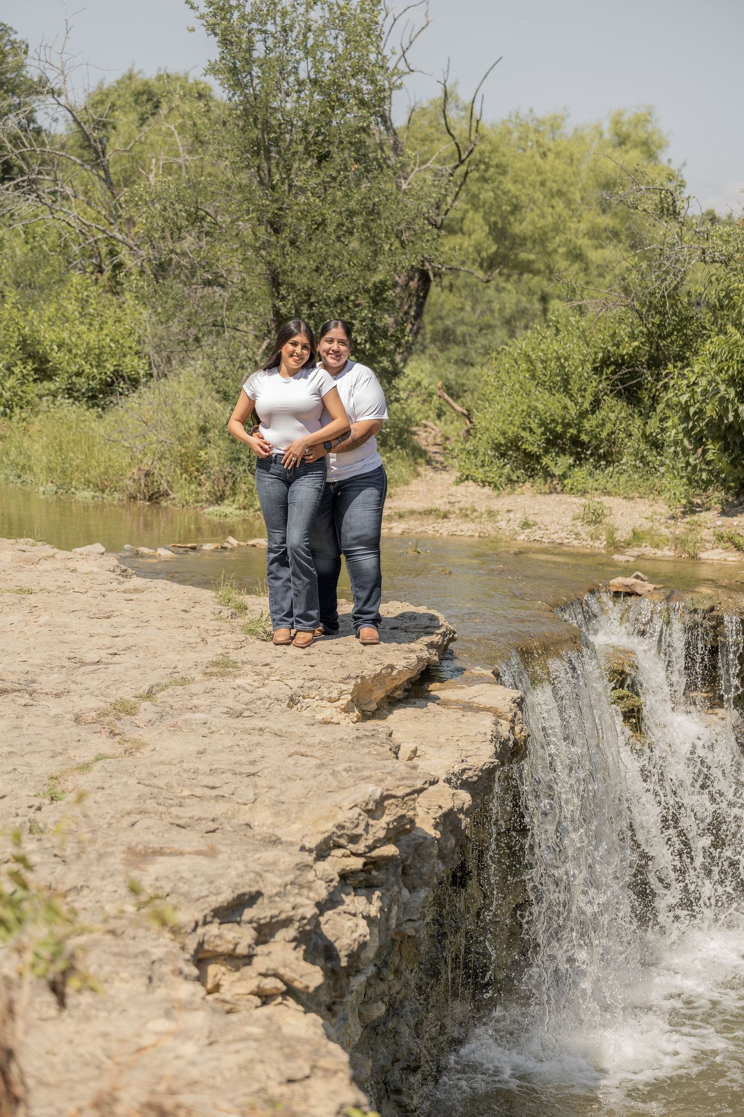 Two women in white tops and jeans smile together on rocks beside a small waterfall in nature.