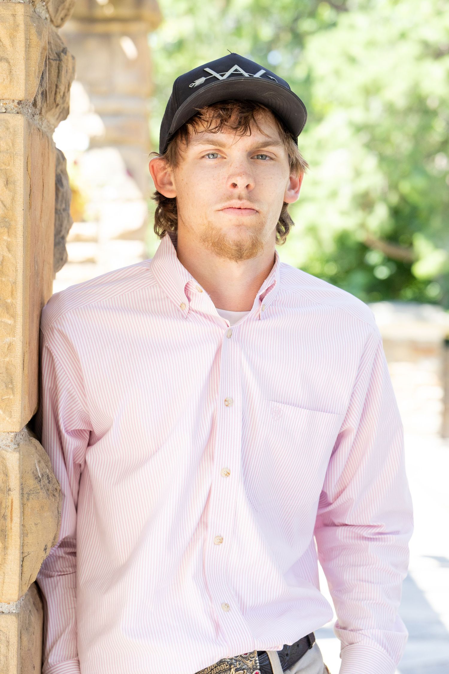 Young man in pink button-up shirt and black cap leaning against stone pillar outdoors.