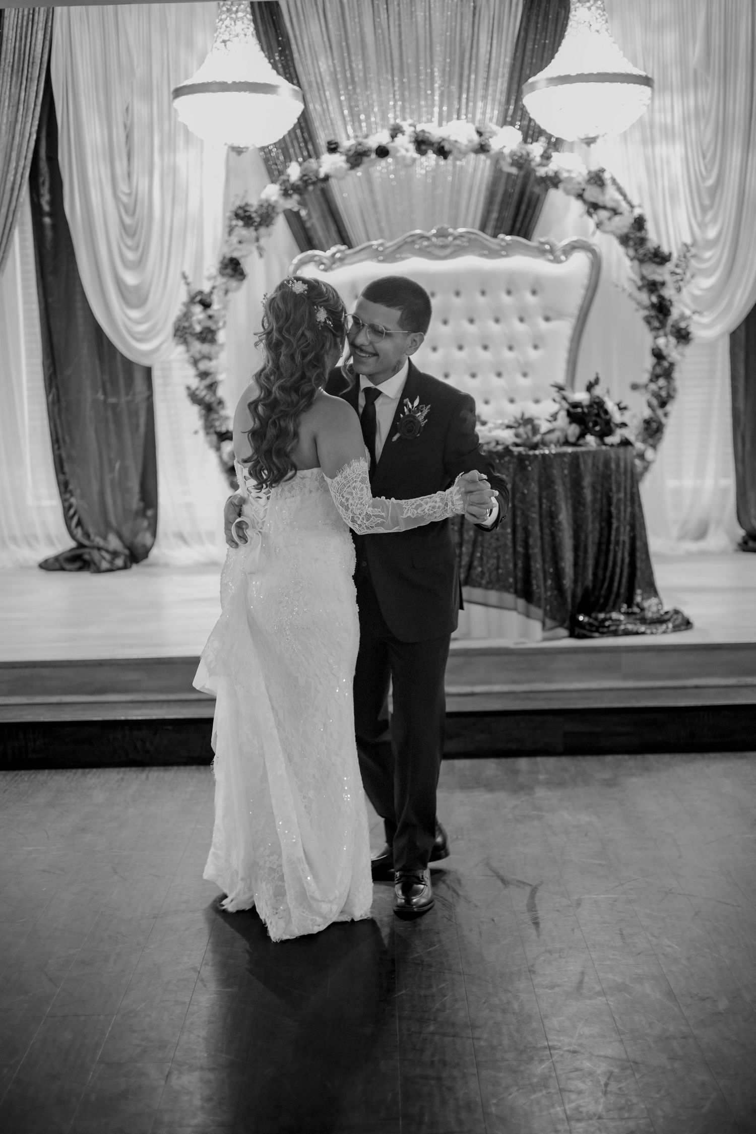 Bride and groom sharing their first dance in elegant ballroom with floral arch backdrop, black and white.