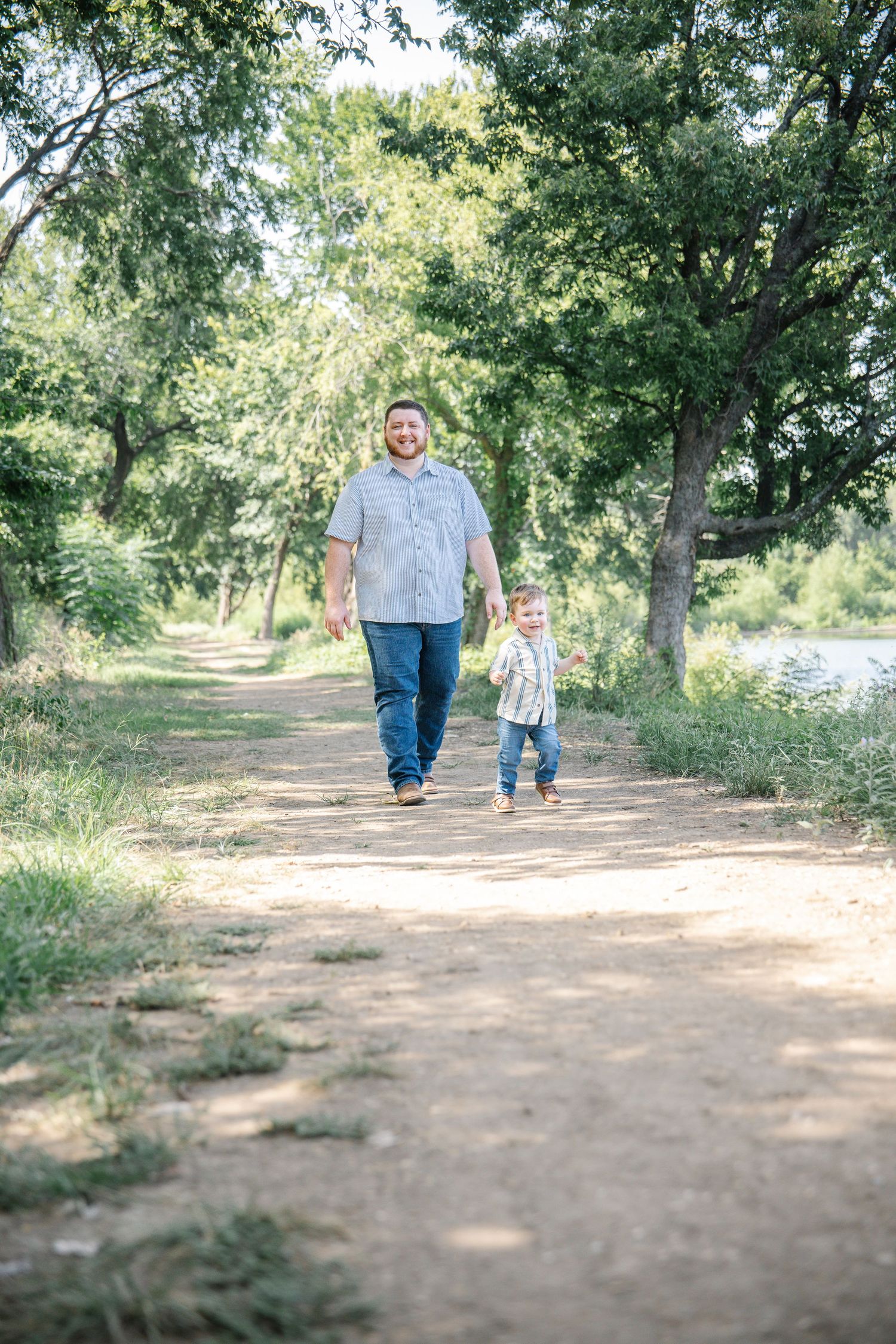 A father and toddler son walk hand-in-hand down a sunny tree-lined dirt path outdoors.