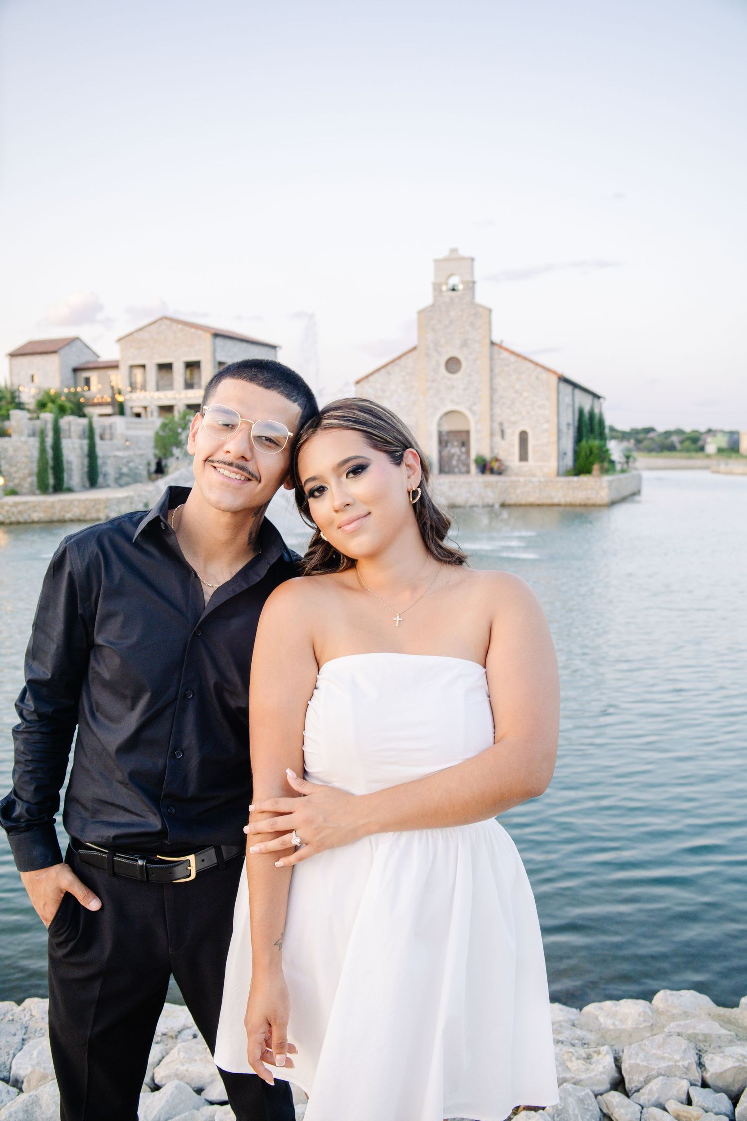 Couple posing by a lakeside stone chapel, man in black shirt and woman in white strapless dress, smiling together.