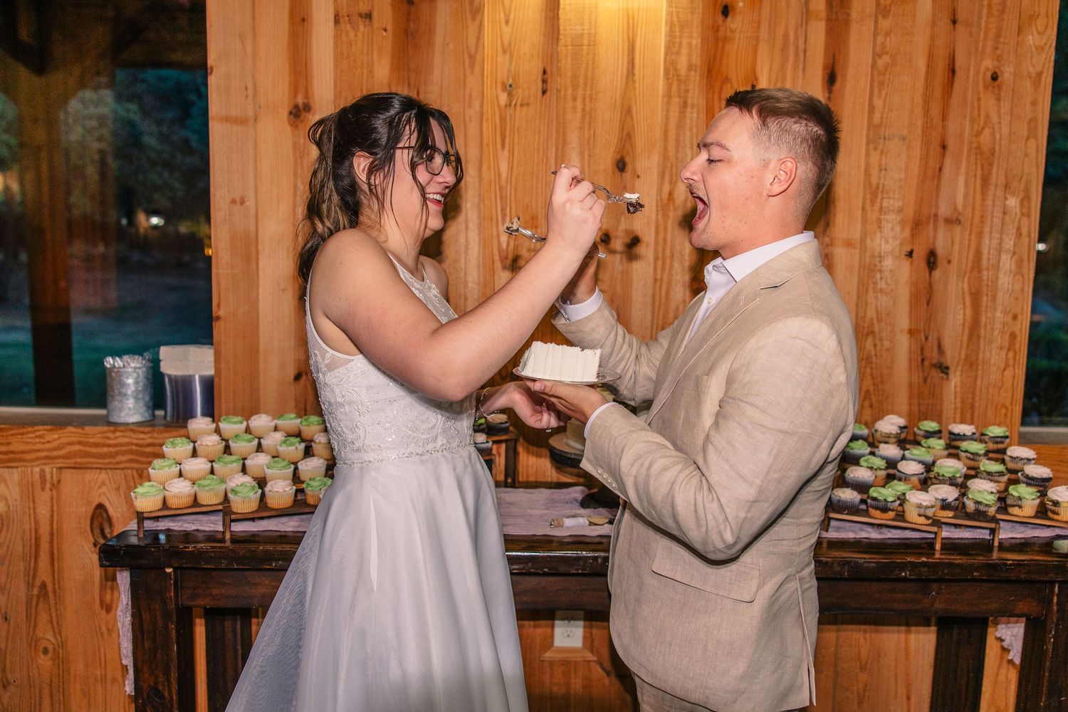 Bride in a white gown and groom in a tan suit playfully feed each other wedding cake at a rustic wooden reception with cupcakes behind them.