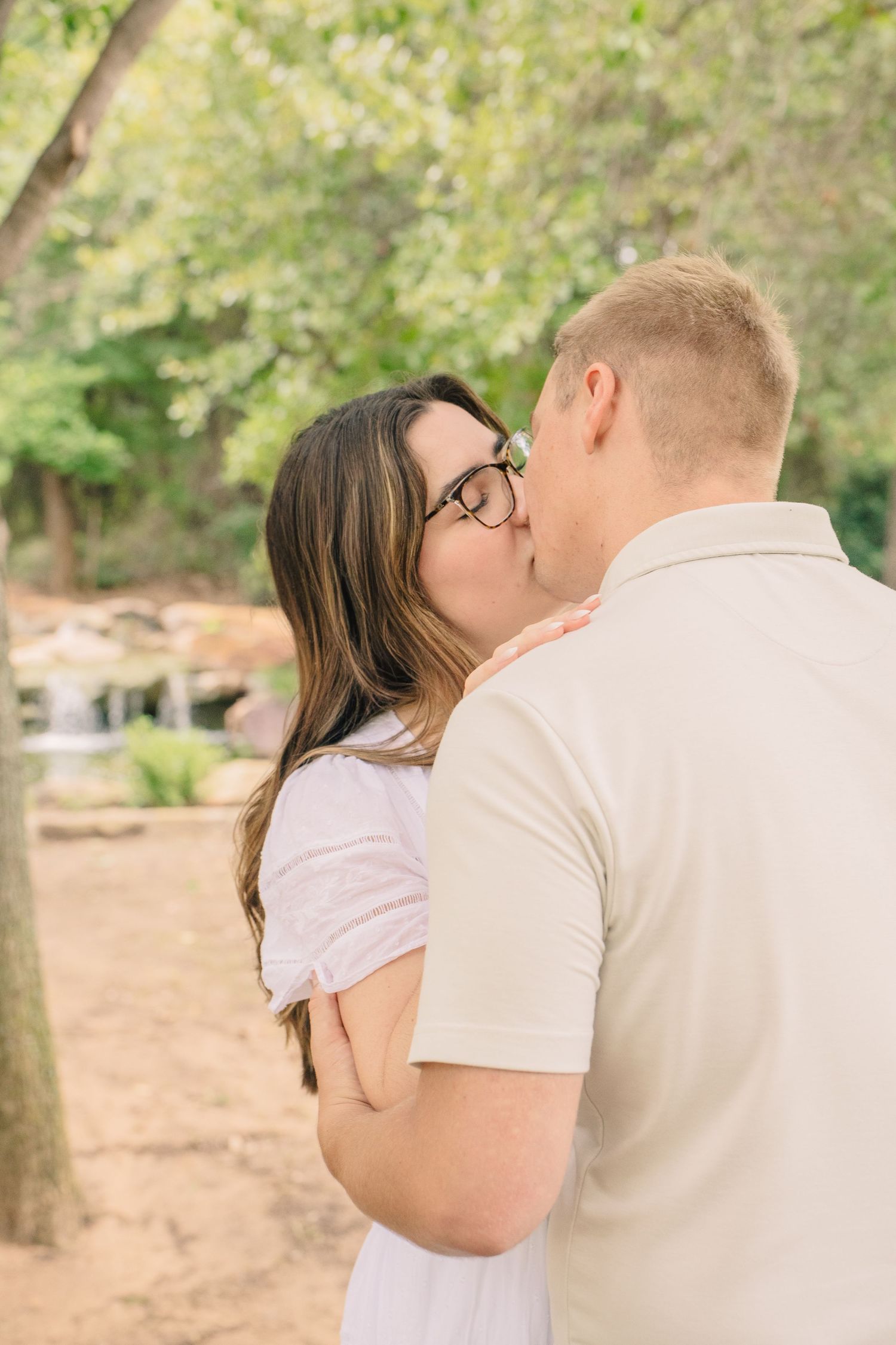 A couple shares a romantic kiss outdoors in a lush green park setting, woman wearing glasses and pink dress.