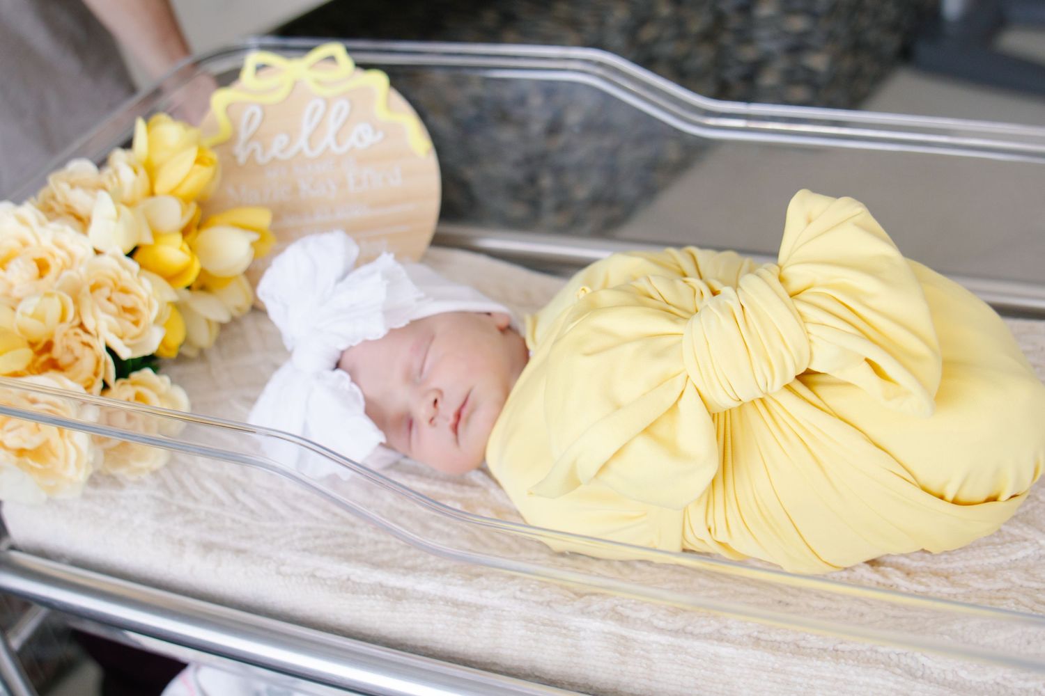 Sleeping newborn baby swaddled in yellow blanket in hospital bassinet with flowers.