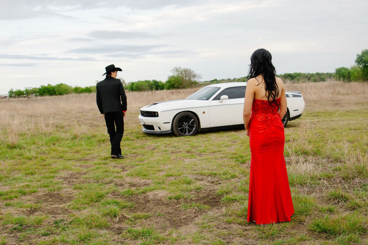 Couple in formal attire standing apart in a field with a white sports car nearby.