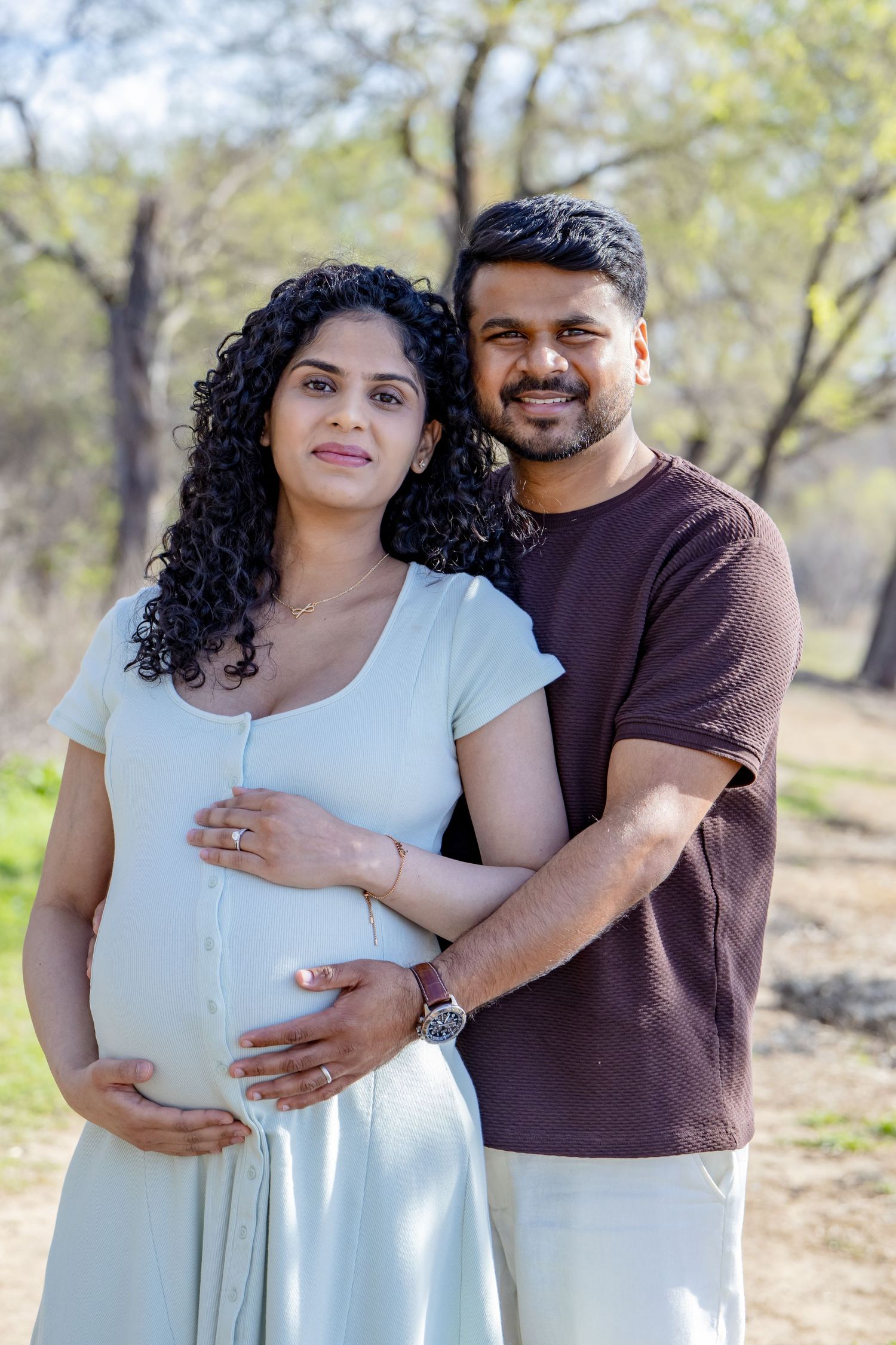 Smiling pregnant Indian couple posing together outdoors, man embracing woman's baby bump in park.