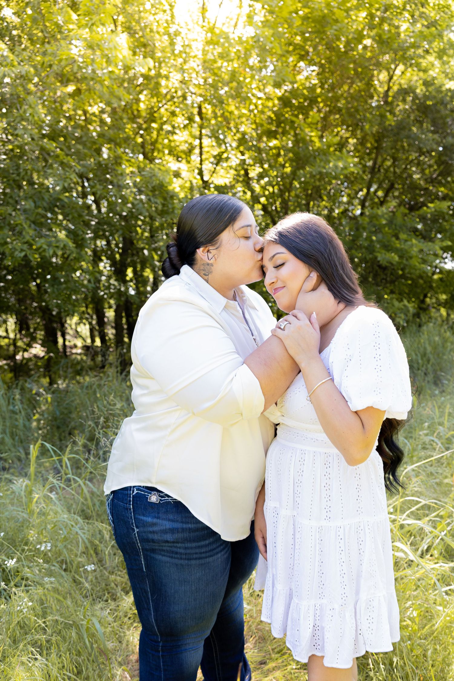Two women share a tender forehead kiss outdoors in a sunlit field surrounded by lush green trees.