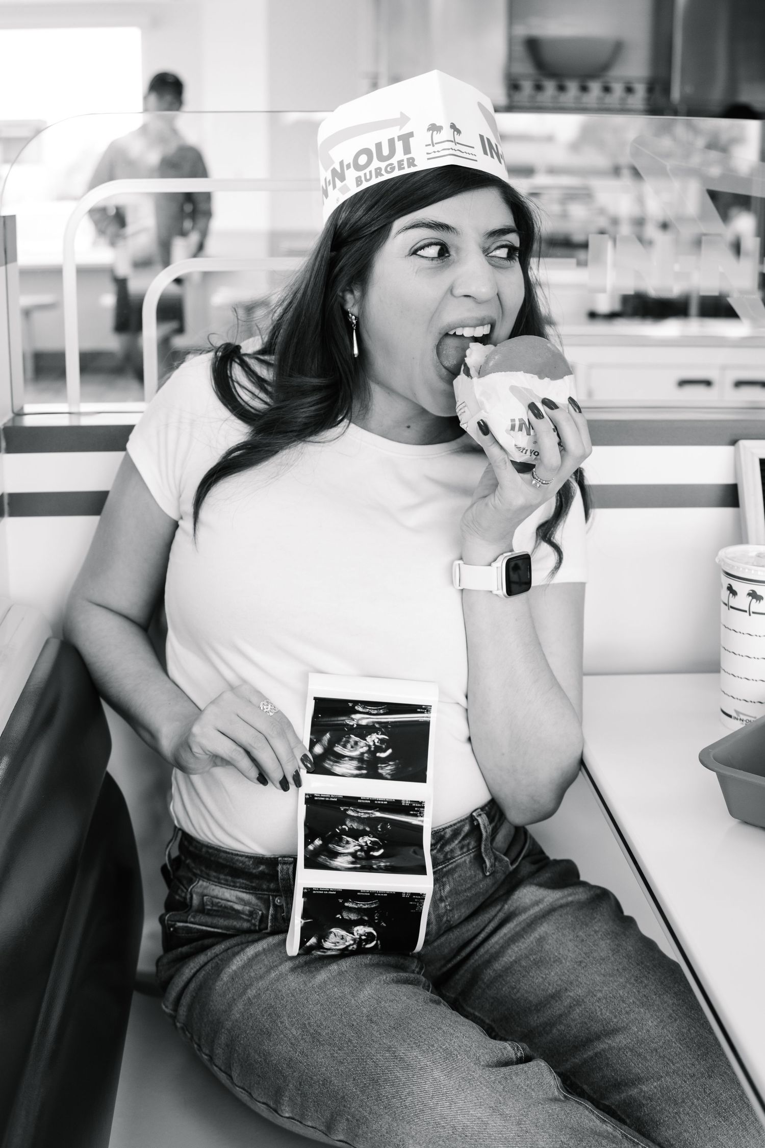 Pregnant woman wearing In-N-Out paper hat eating a burger while holding ultrasound photos in a restaurant booth.