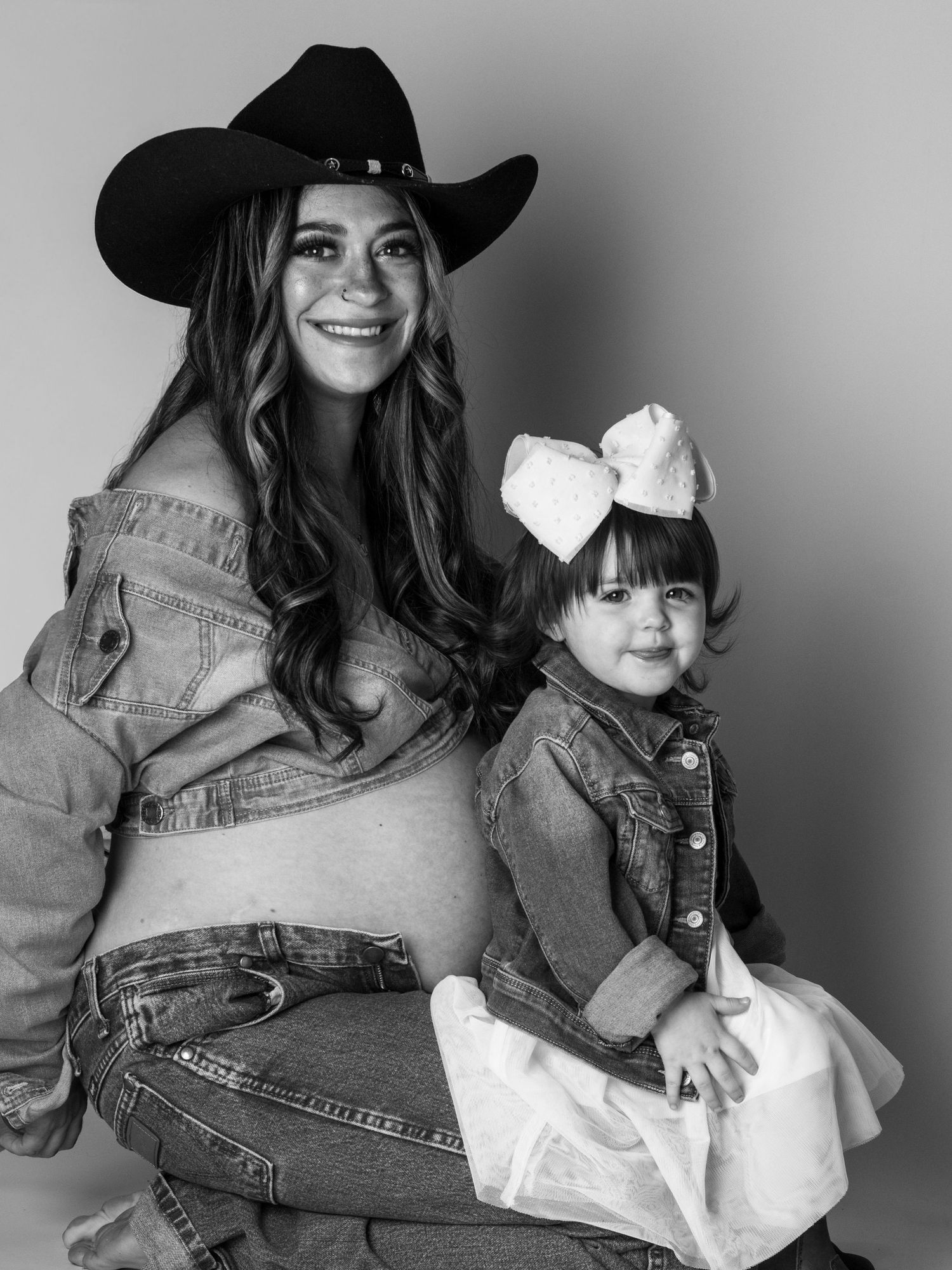 Black and white maternity photo of a smiling woman in cowboy hat and denim with a toddler girl wearing a bow.
