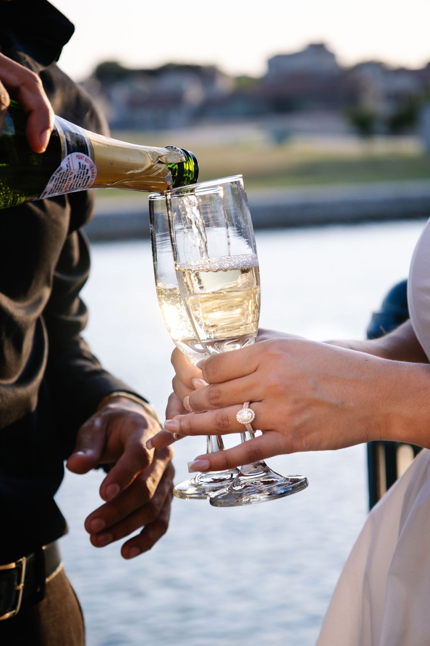 Champagne being poured into crystal flutes held by a bride wearing an engagement ring near a river.