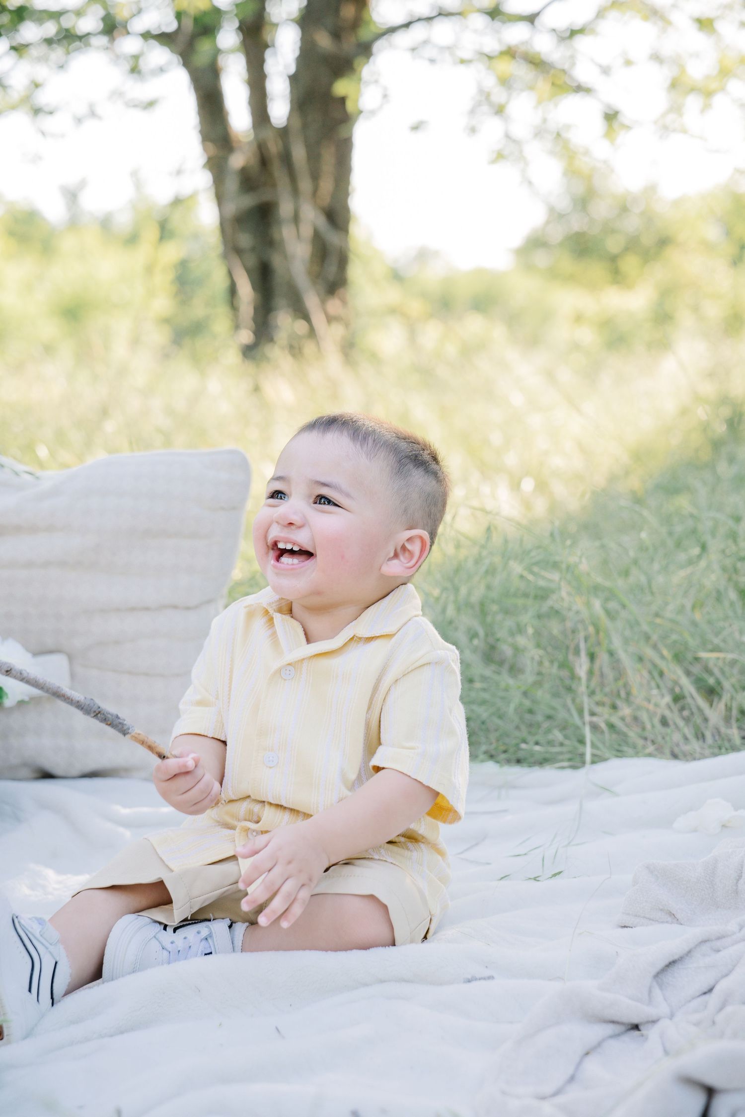A laughing toddler boy in a yellow outfit sits on a blanket outdoors, holding a stick near a tree.