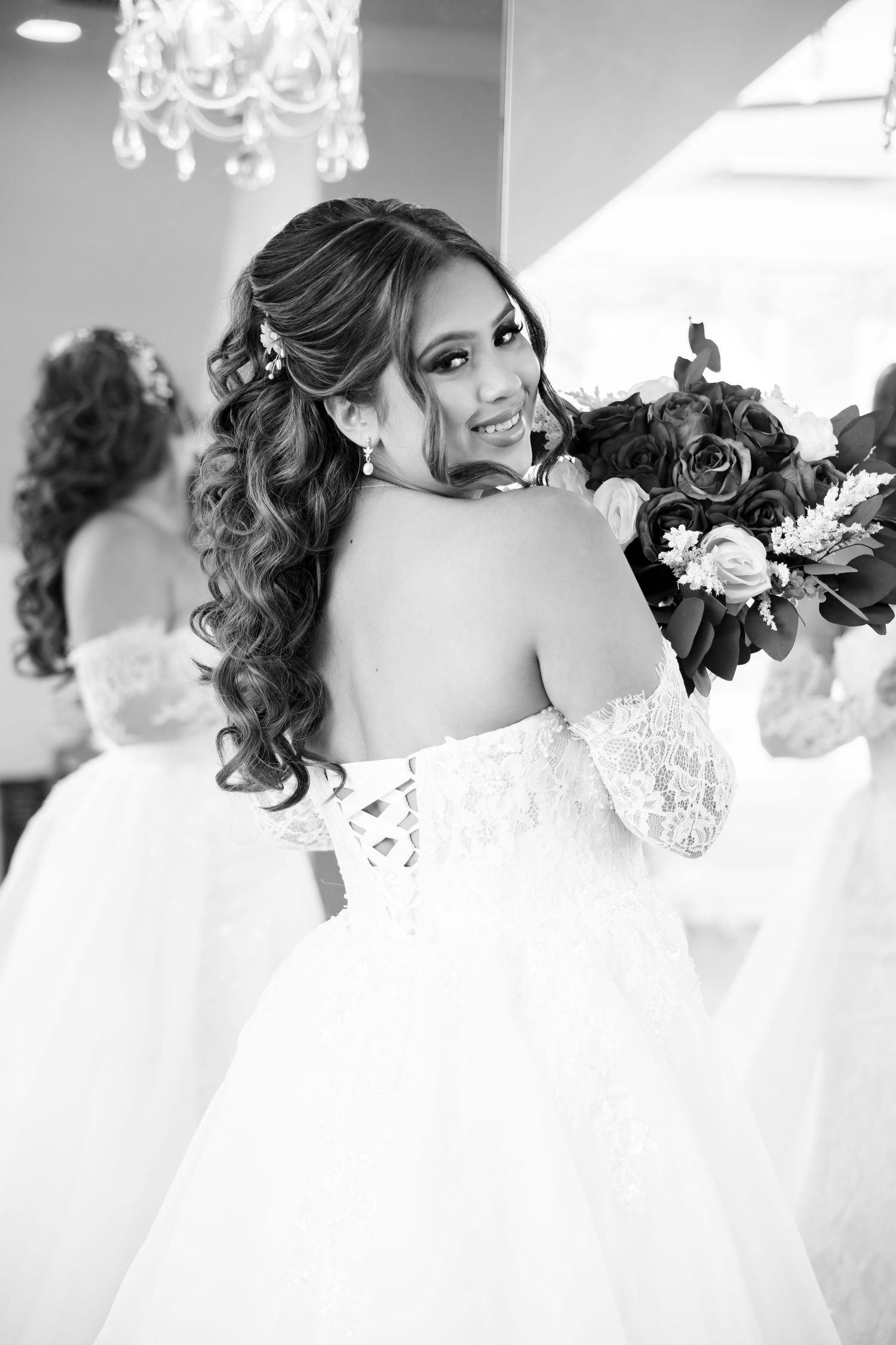 Bride in off-shoulder lace wedding gown holding bouquet, smiling over shoulder in black and white photo.