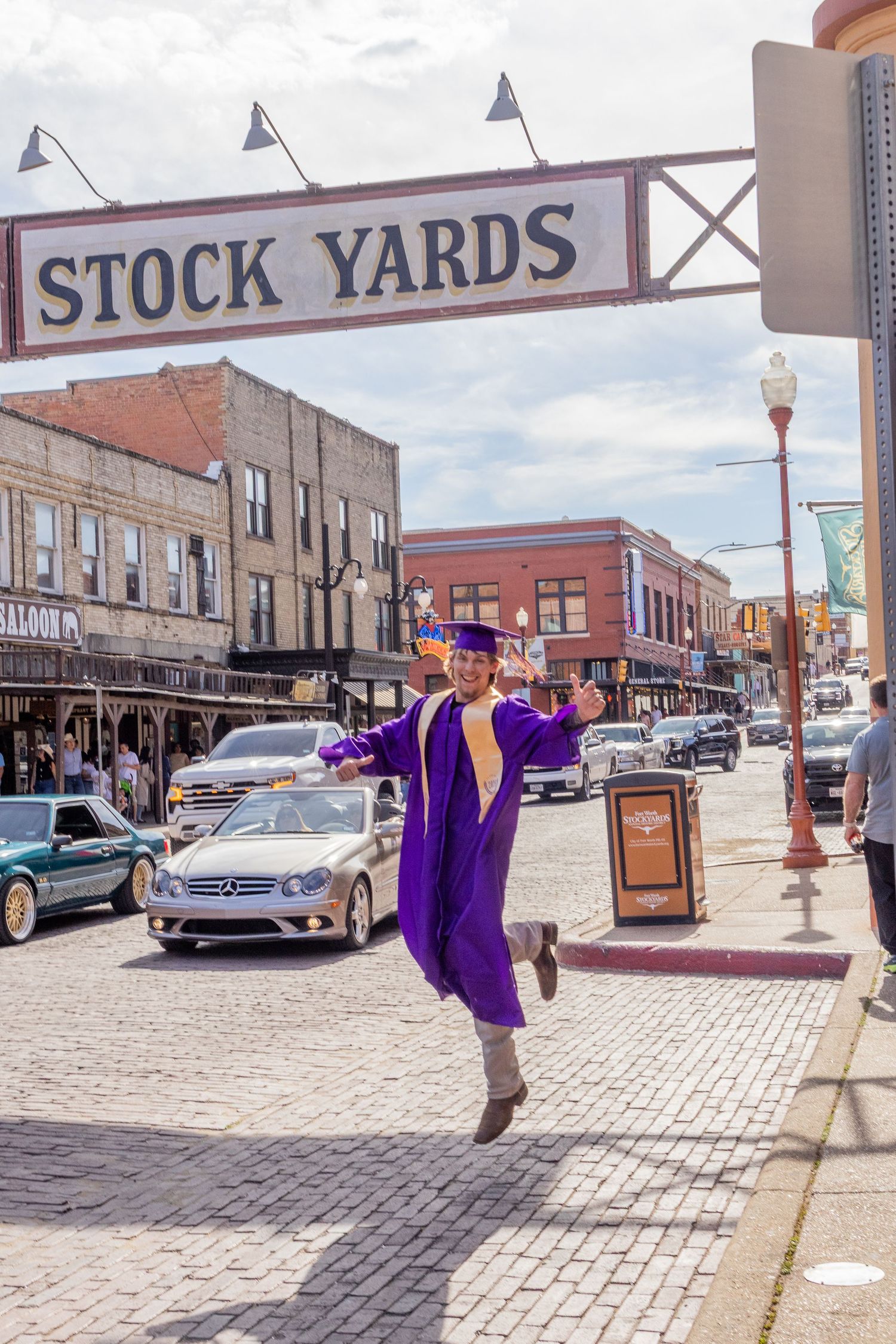 Excited graduate in purple cap and gown jumps joyfully under Fort Worth Stock Yards sign.