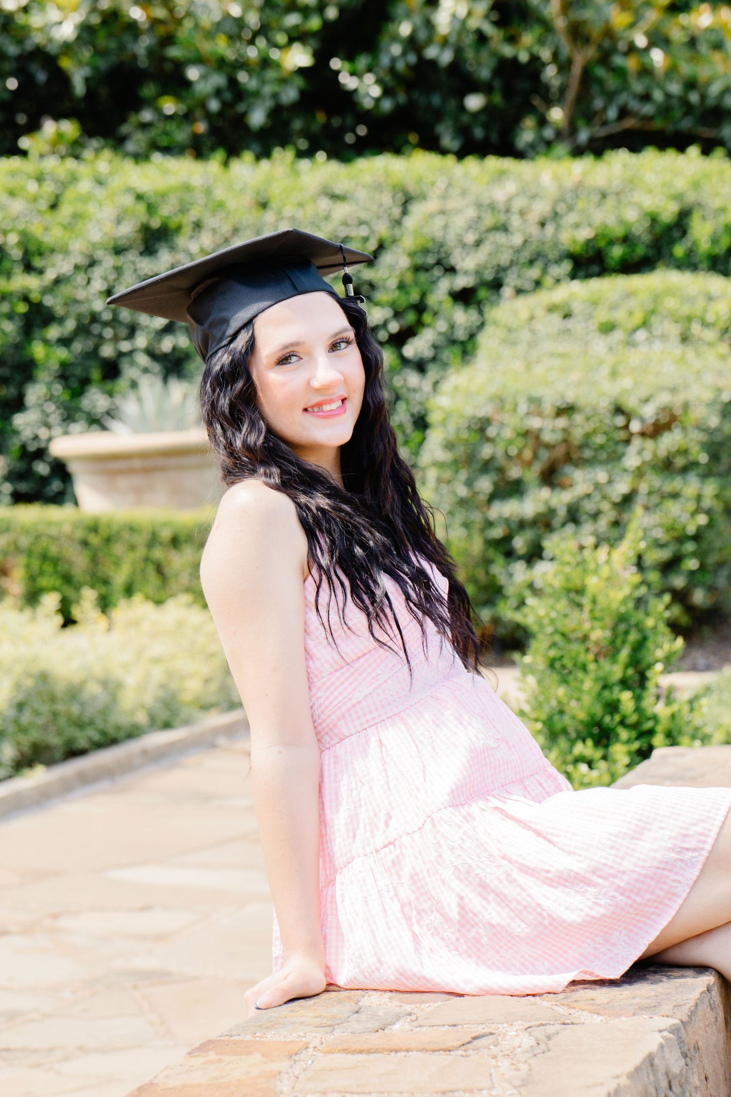 A young woman in a pink dress and graduation cap sits on a stone wall smiling in a garden.