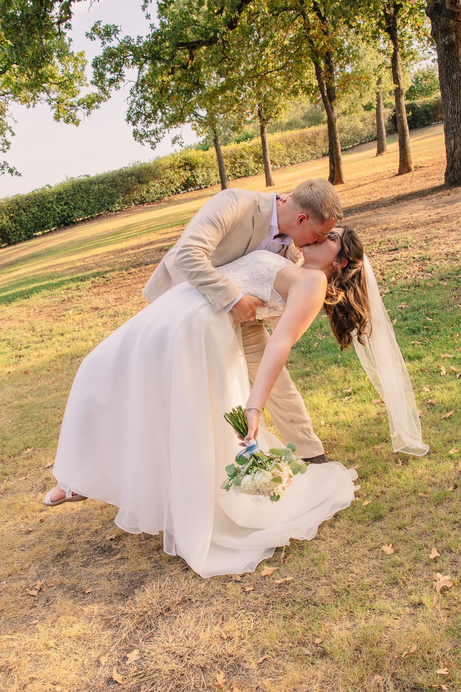 Groom dips and kisses his bride, who holds a bouquet and wears a veil, on a sunlit grassy lawn at an outdoor wedding.