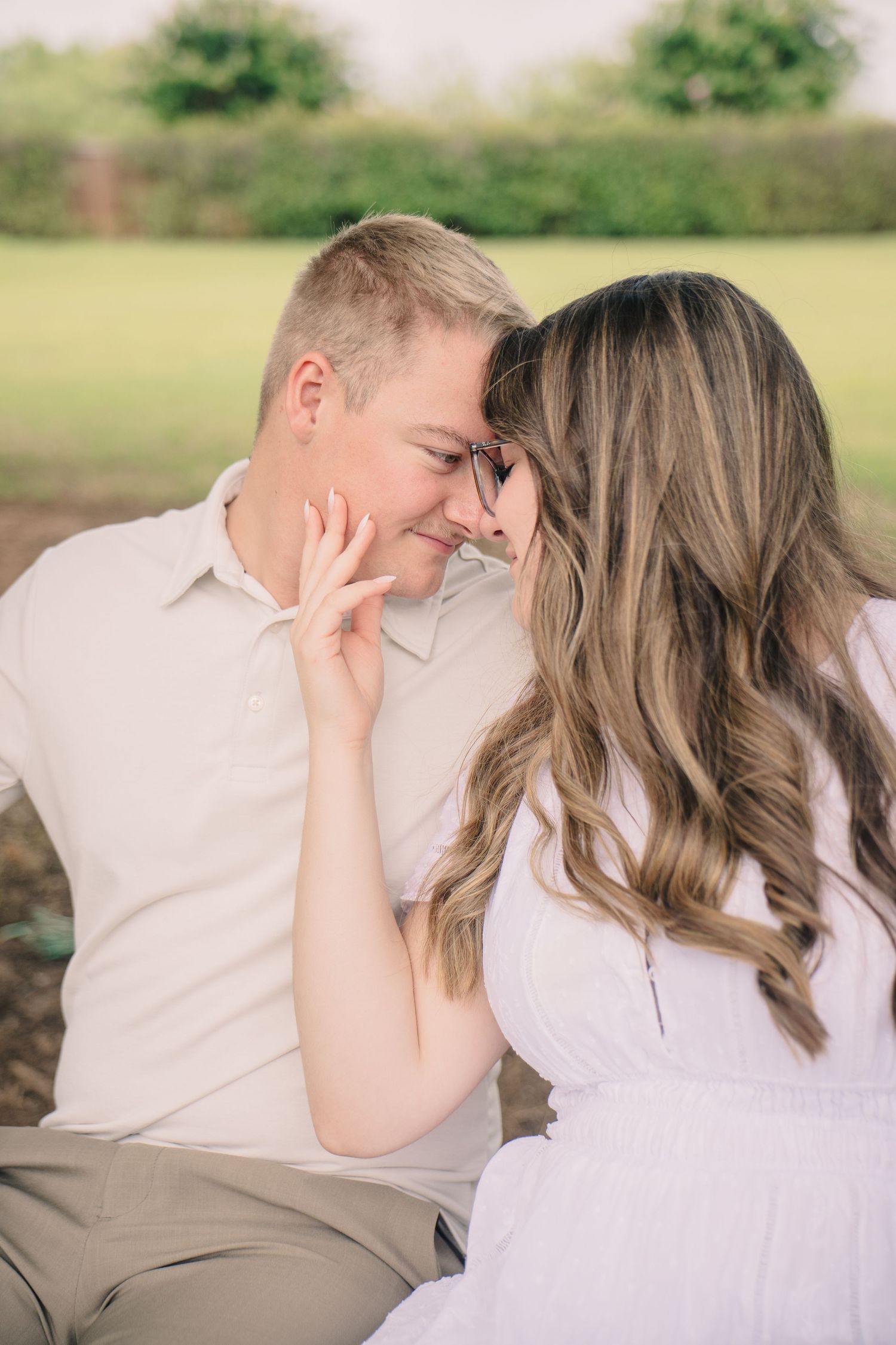Couple sharing a romantic moment outdoors, foreheads touching and smiling, woman in white dress with wavy hair, green field background.