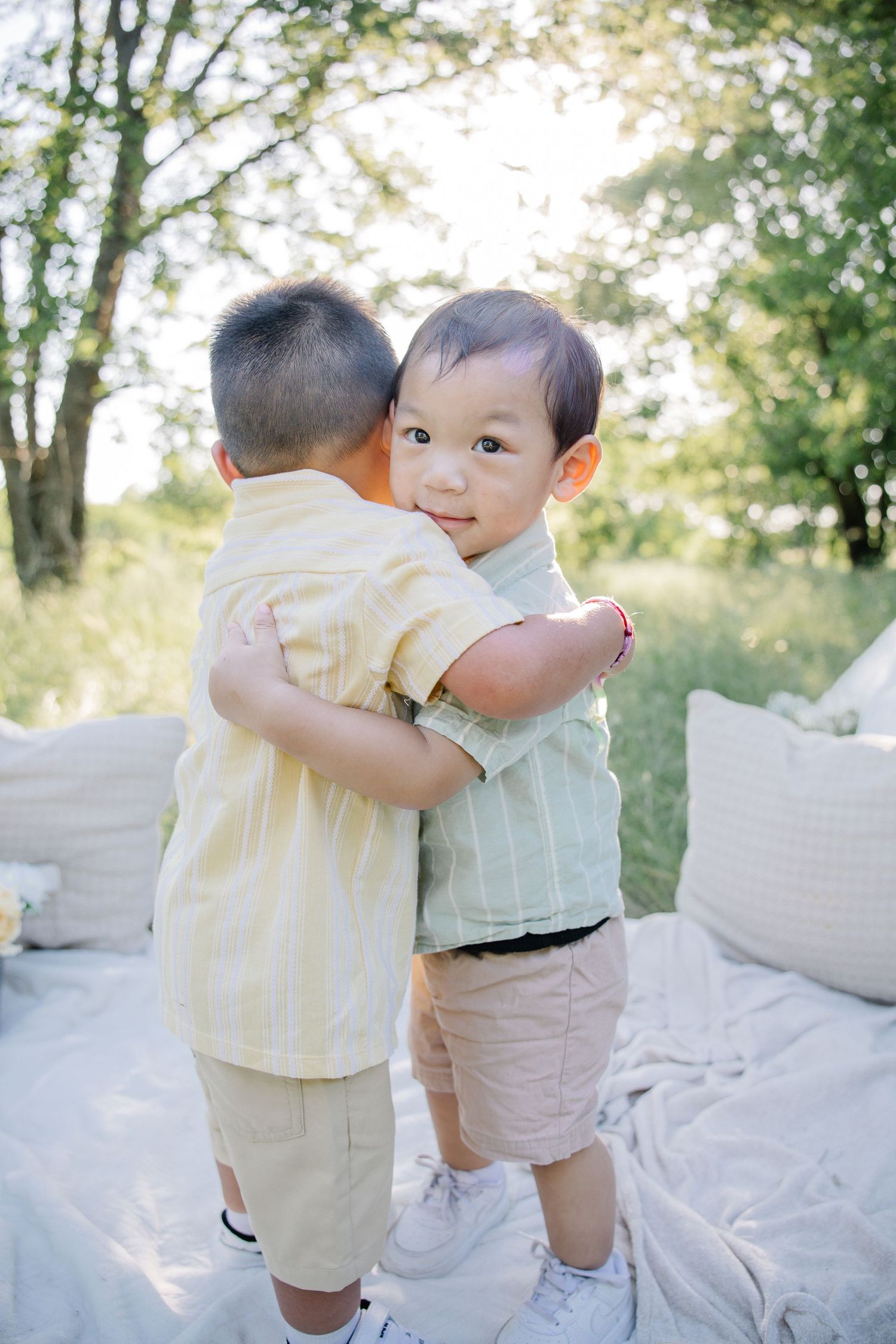 Two young Asian boys hugging outdoors on a blanket, surrounded by trees in soft natural light.