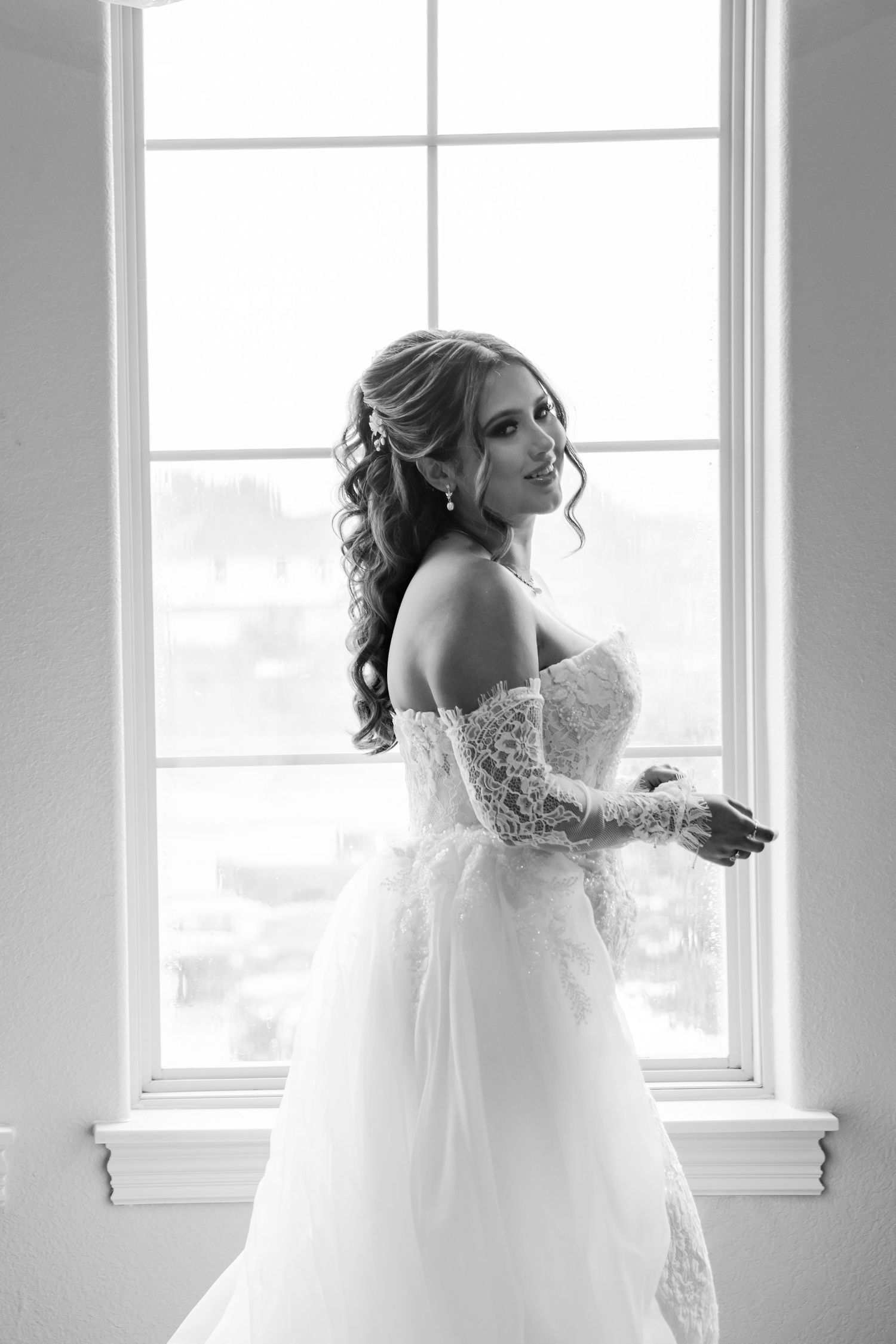 Elegant bride in lace off-shoulder wedding gown poses by large window in black and white photo.