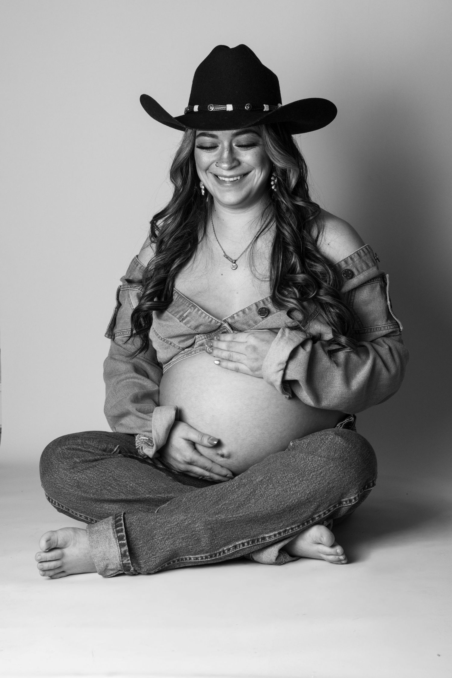 Black and white maternity photo of smiling pregnant woman in cowboy hat, jeans, and denim jacket sitting cross-legged.