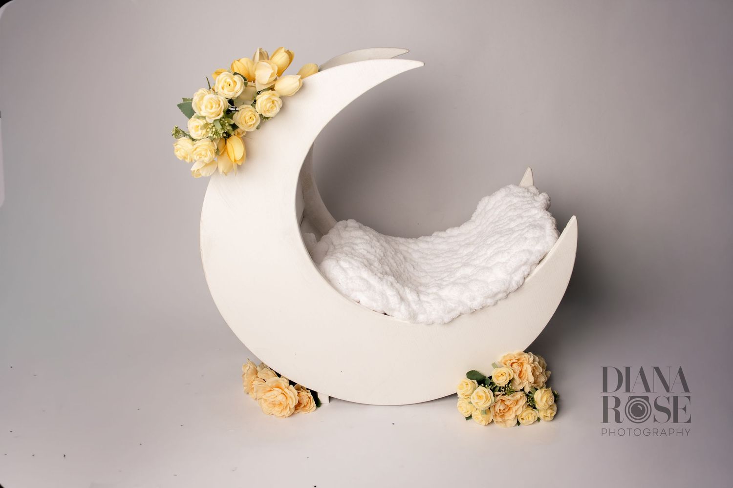 White crescent moon newborn photography prop decorated with yellow roses on a gray studio background.