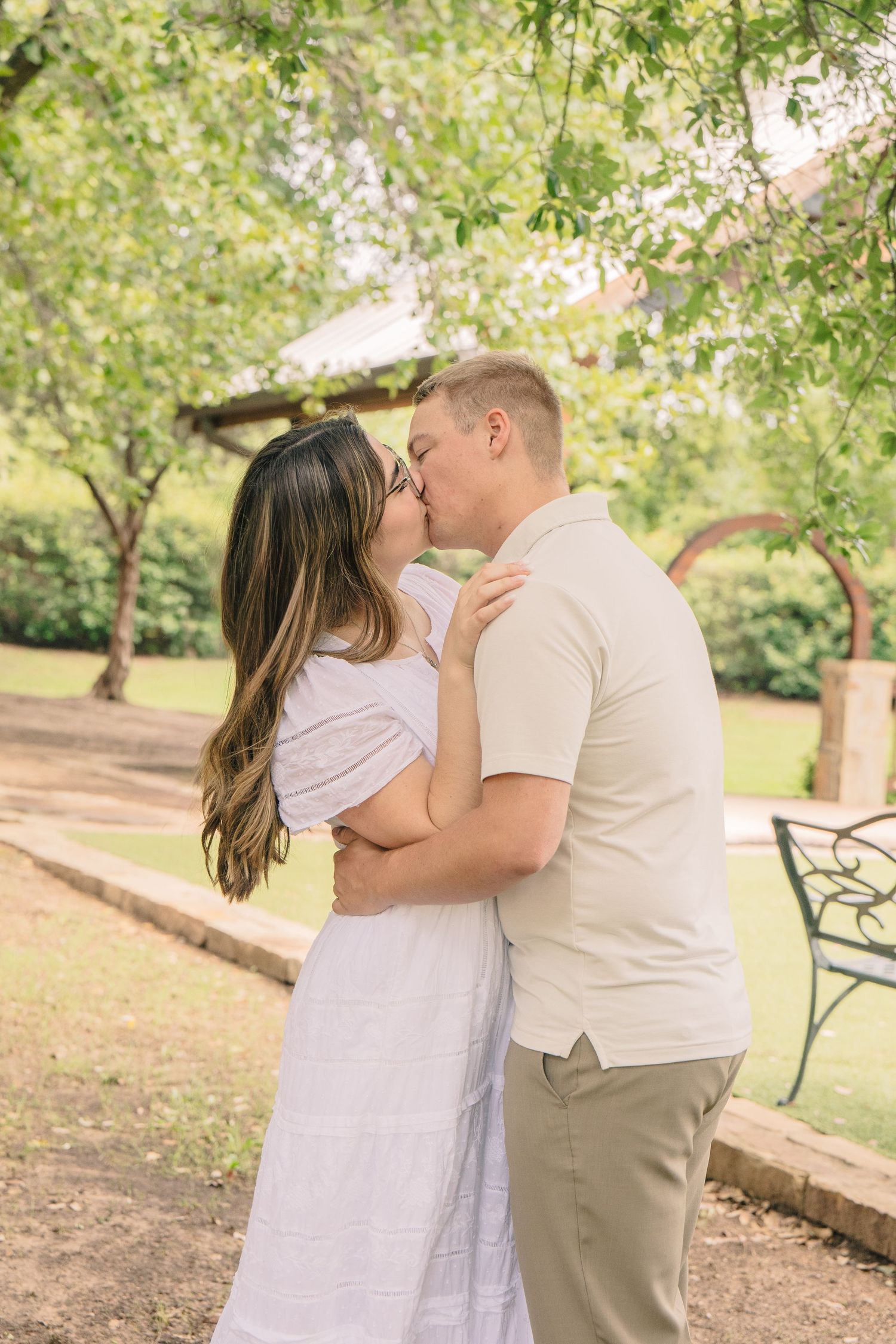 A couple shares a romantic kiss in a lush green park, woman in white dress, man in beige outfit.