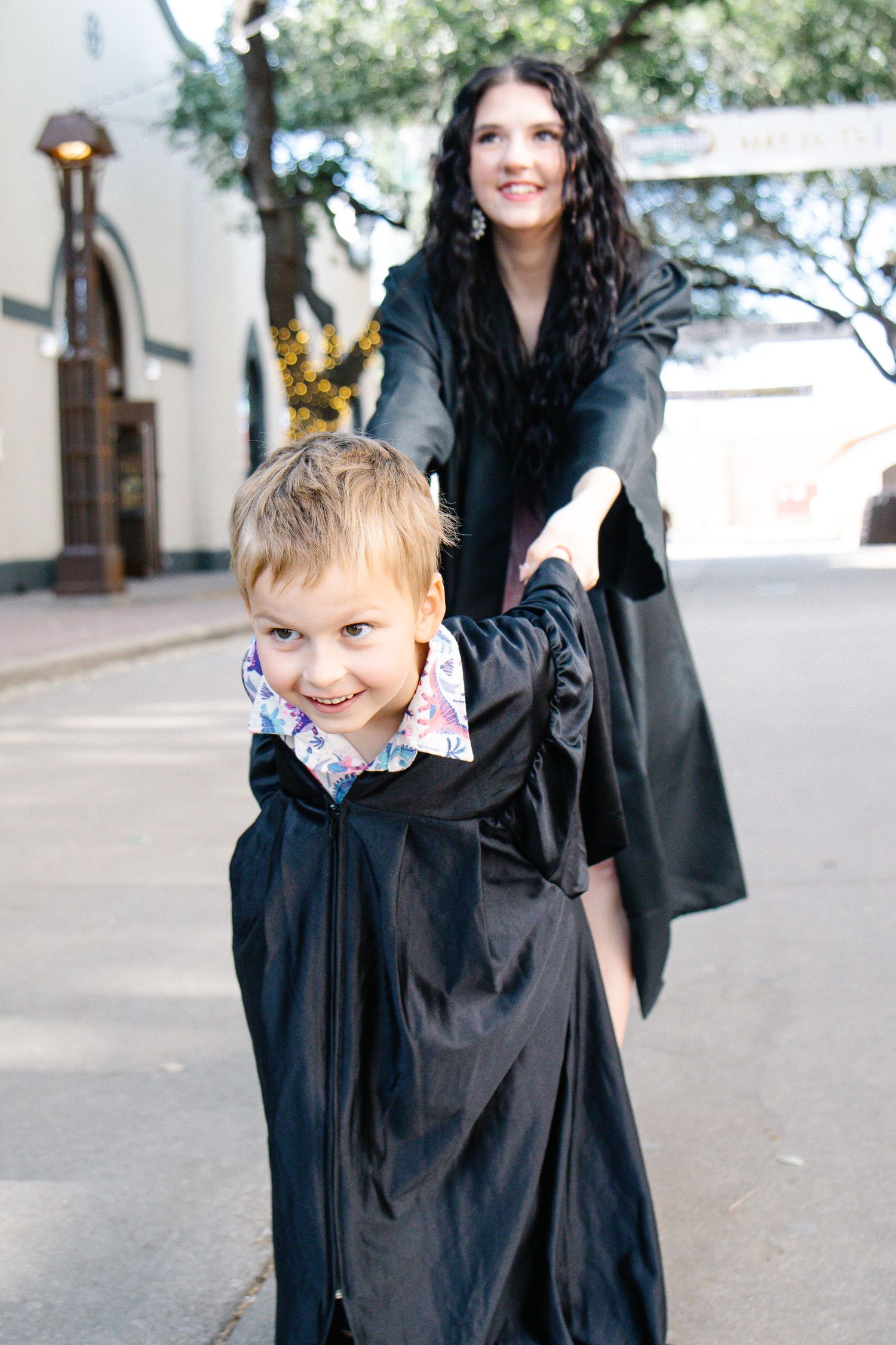 Smiling woman in graduation gown holds hands with young boy wearing oversized gown outdoors.
