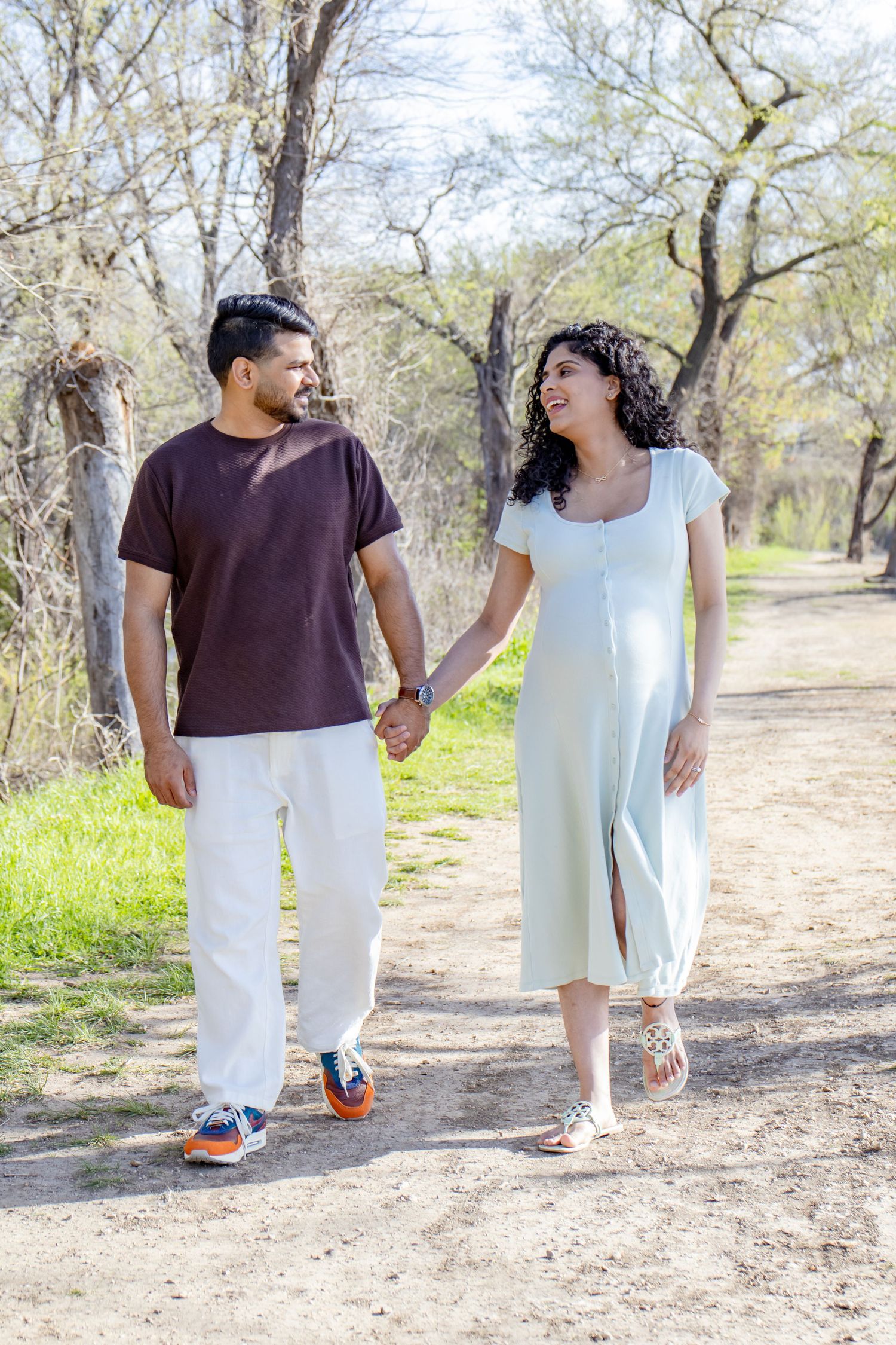 Happy expecting couple holding hands walking on a sunny nature trail surrounded by bare trees.