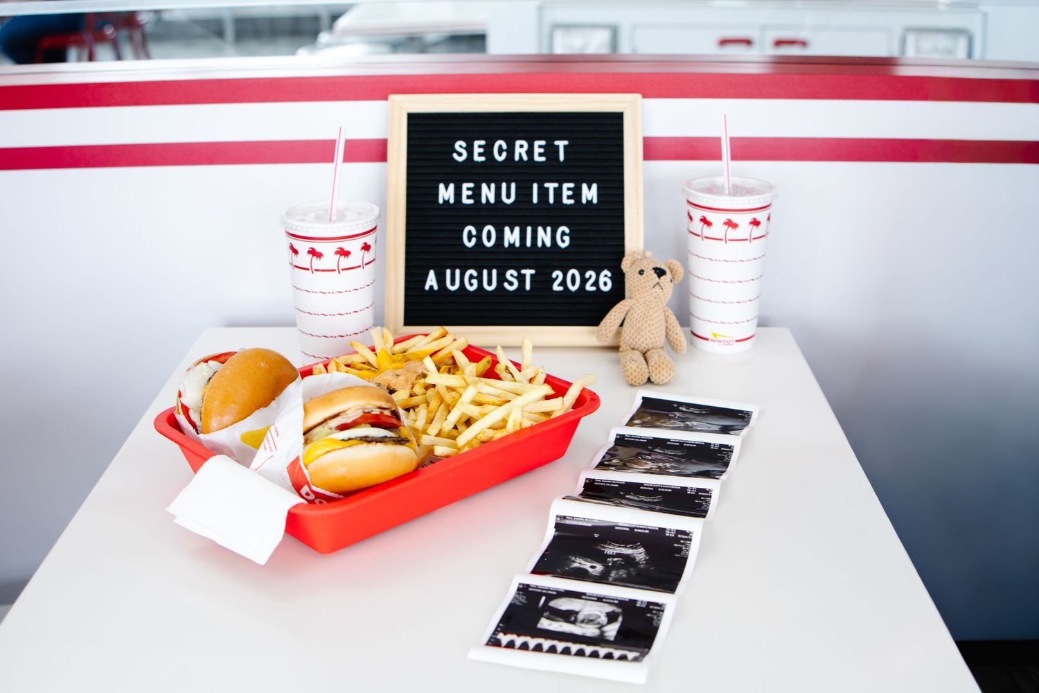 In-N-Out Burger meal with ultrasound photos, a teddy bear, and a letter board announcing a baby coming August 2026.