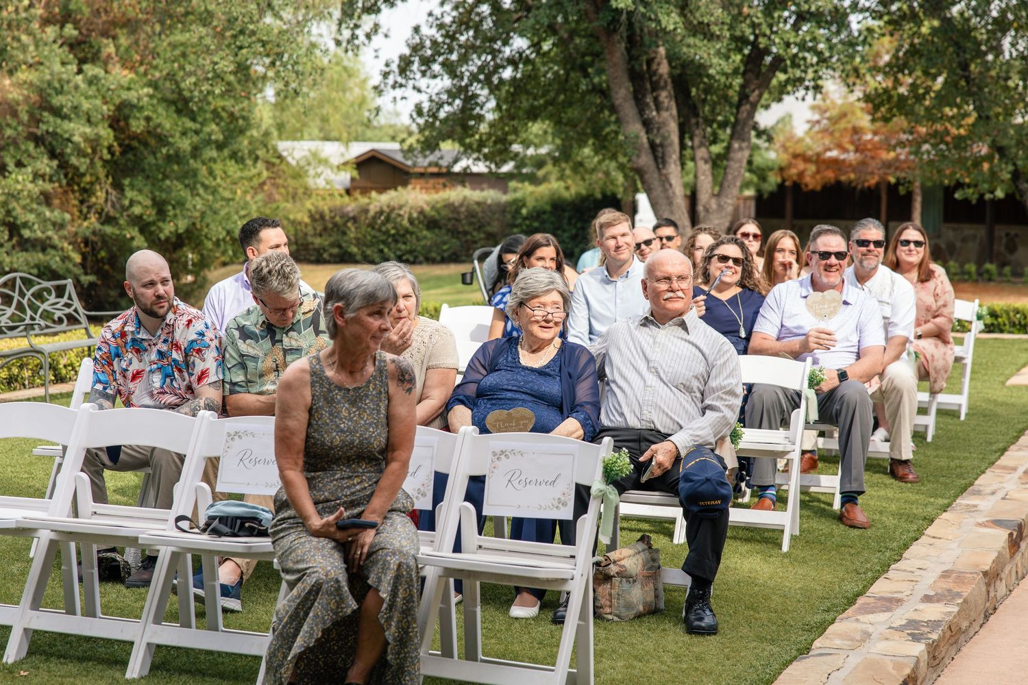 Wedding guests sit on white folding chairs in an outdoor garden ceremony, surrounded by green trees and grass.
