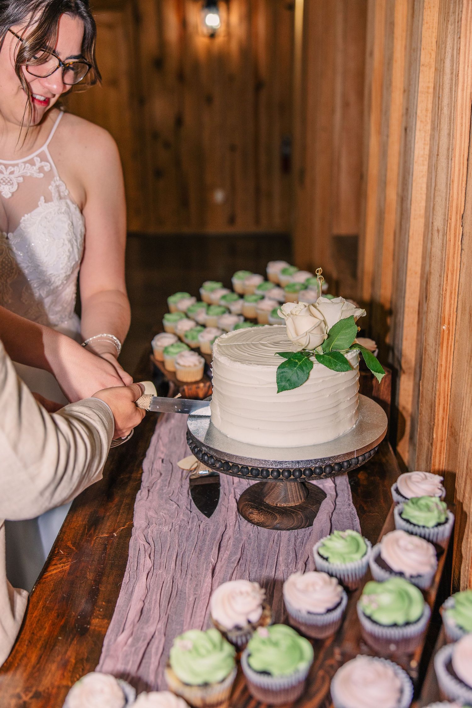 A couple cuts a white wedding cake with a rose on top, surrounded by green and pink cupcakes on a rustic table.