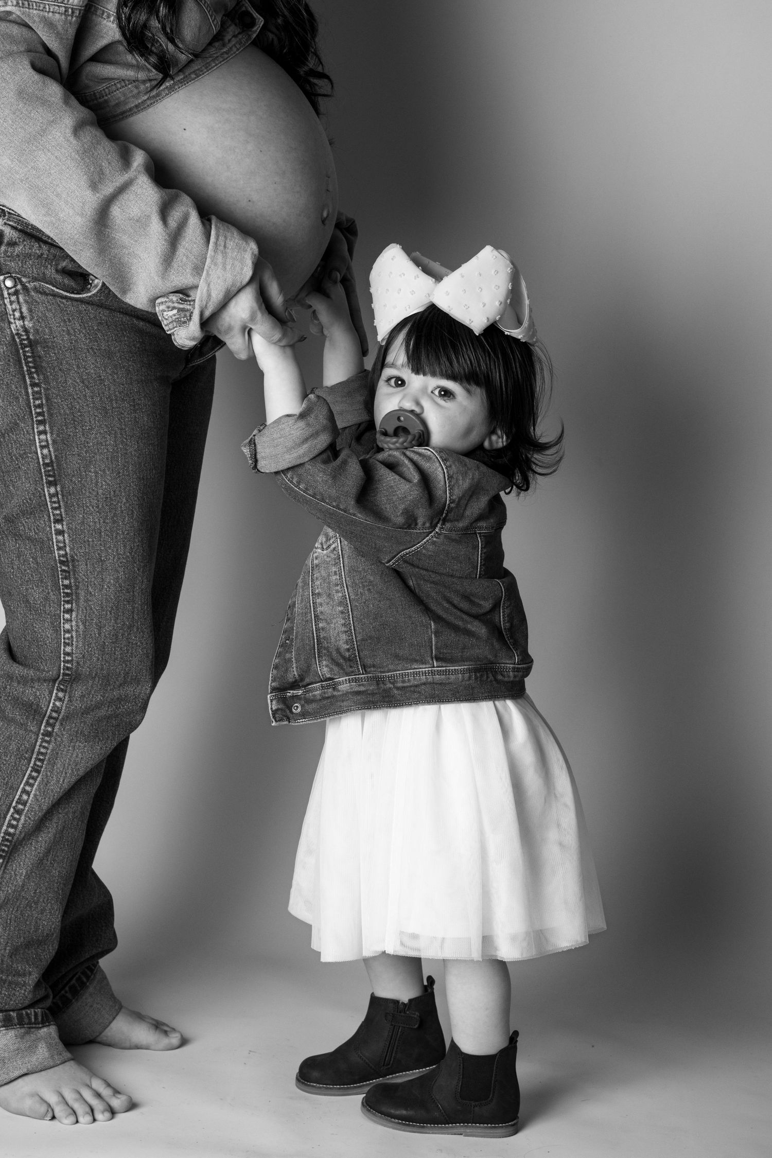 Black and white photo of a toddler girl in denim jacket and white dress holding her pregnant mother's hand.